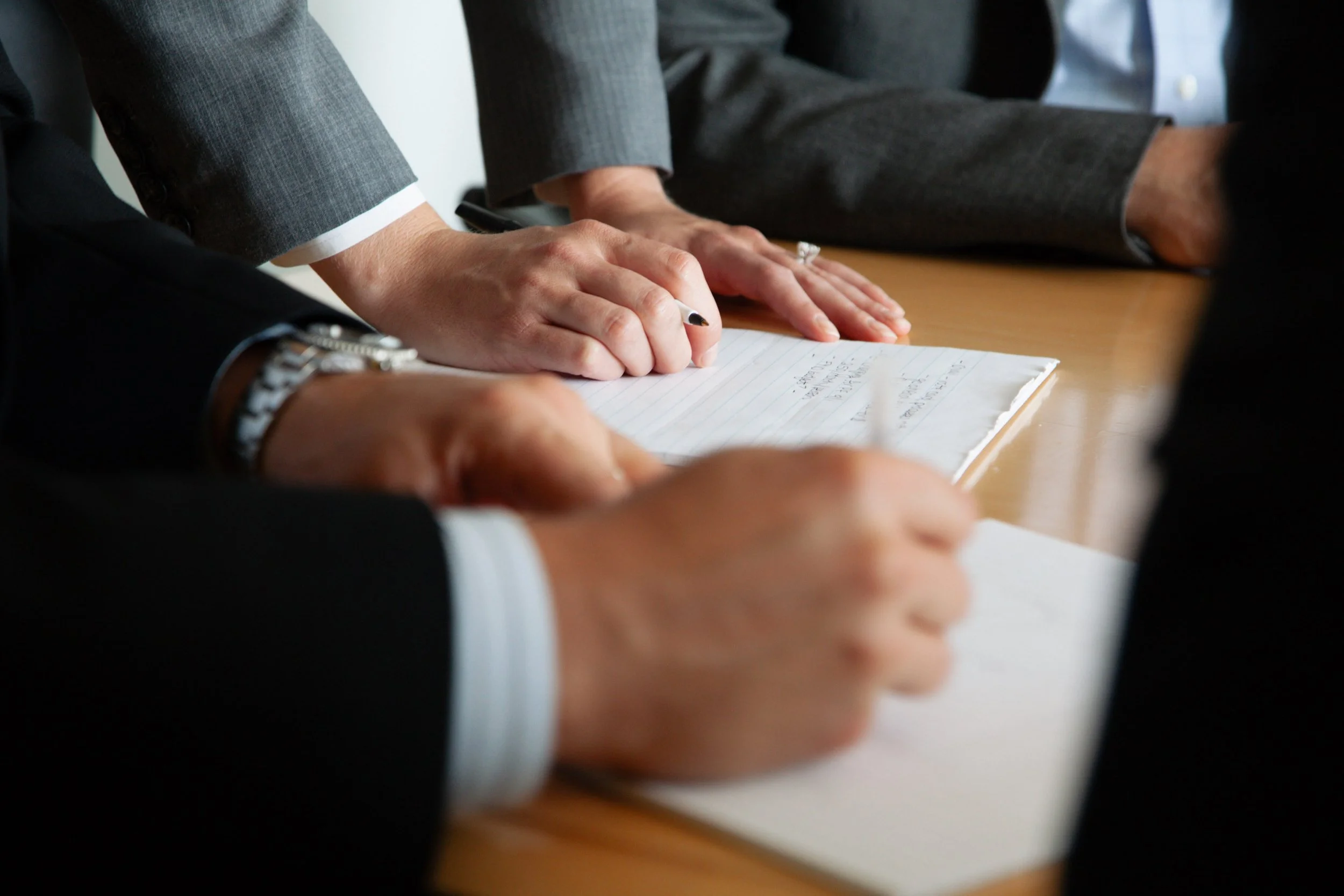 People in business suits participating in a meeting, with one person writing notes on a notepad.