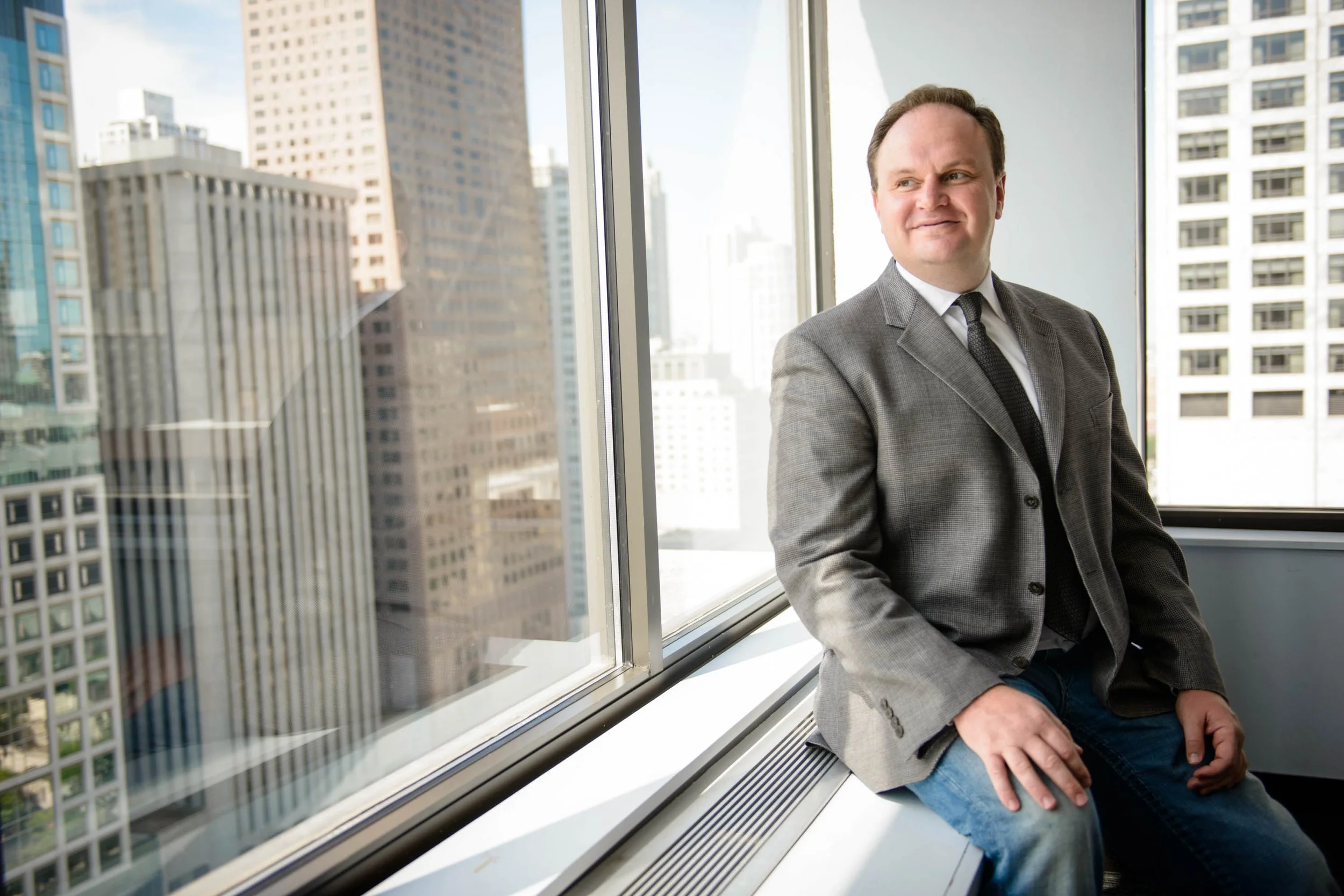 A man in a gray suit sitting on a bench near large windows with a cityscape of tall buildings outside.