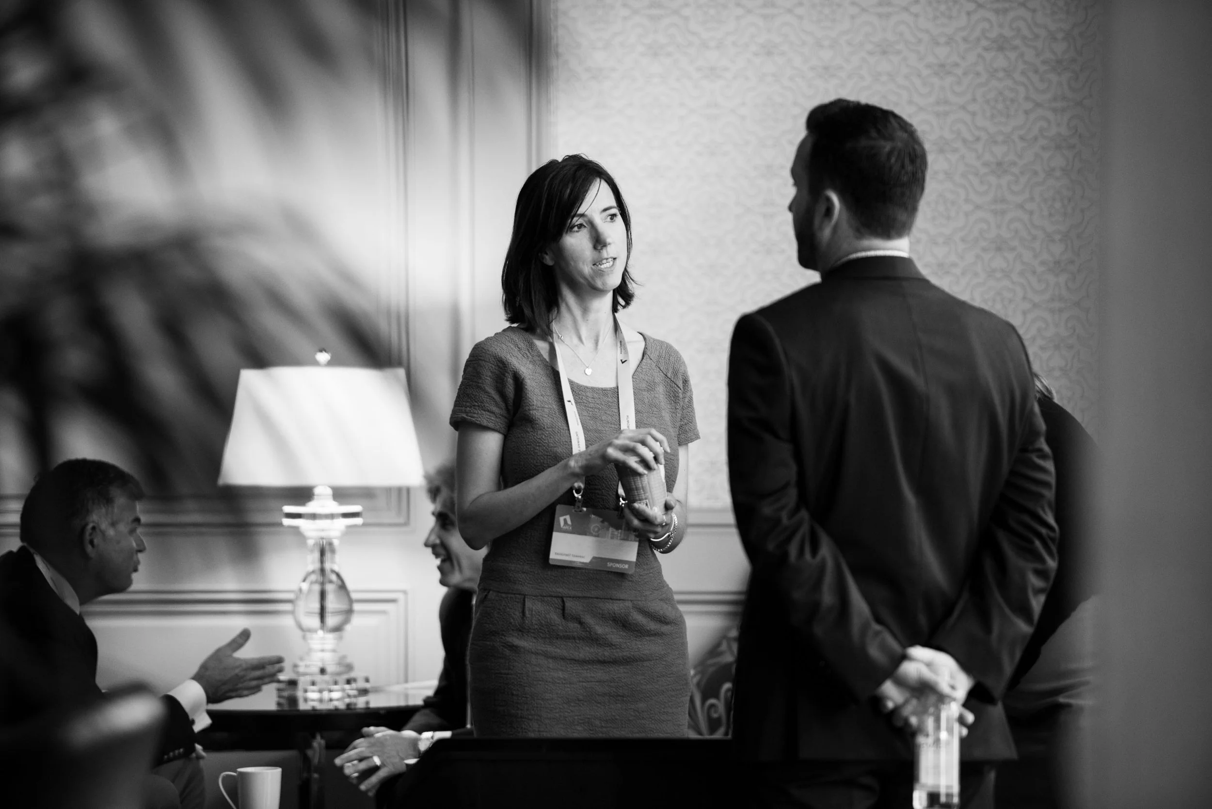 A woman and a man are engaging in a conversation at a professional event. The woman is holding a cup, wearing a name badge lanyard, and is talking to the man dressed in a suit. There are other people around, some sitting at a table with a lamp and a 