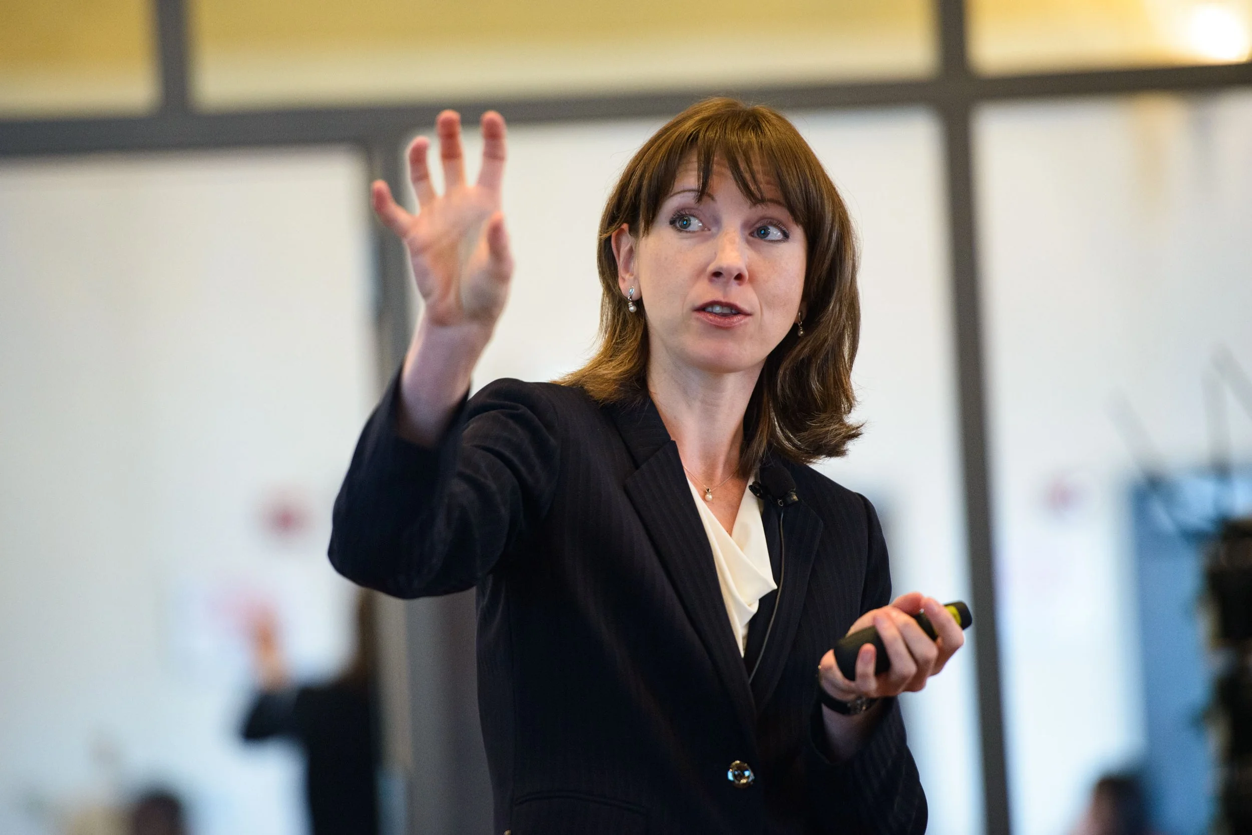 A woman with brown hair and blue eyes, dressed in a dark blazer and white blouse, is speaking and gesturing with her right hand while holding a device in her left hand at an indoor event or conference.