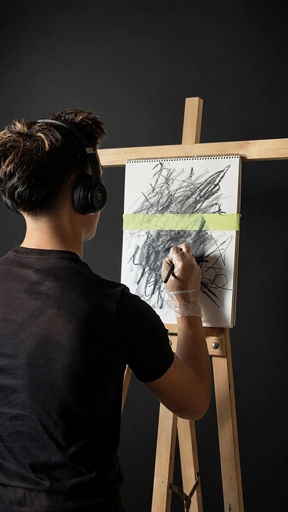 A person wearing headphones and a glove is drawing on a white paper mounted on an easel, with black scribbles and lines, against a dark background.