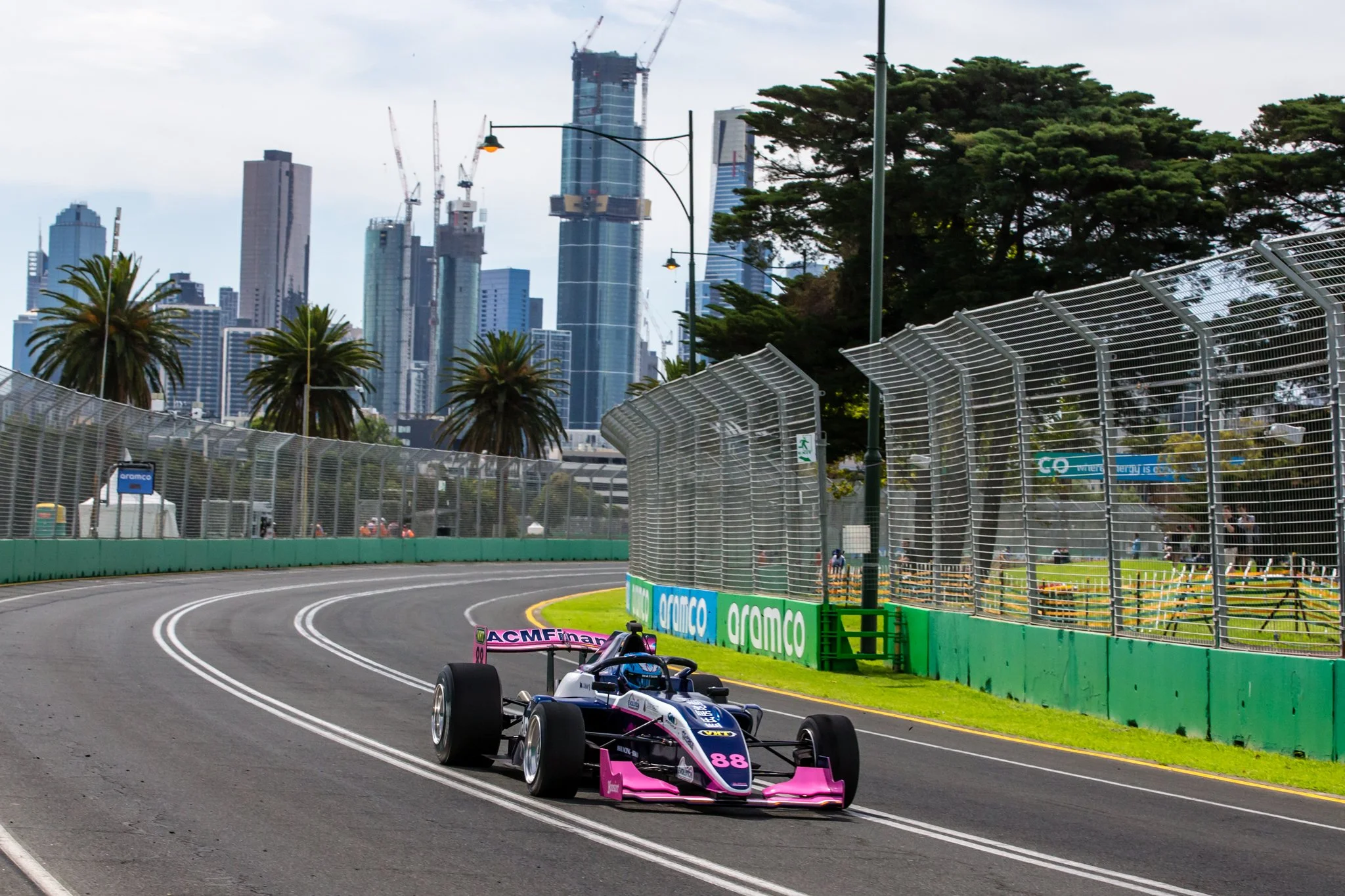 A racecar on a curved race track with the city skyline and tall buildings under construction in the background. The car has pink and black coloring with the number 88.