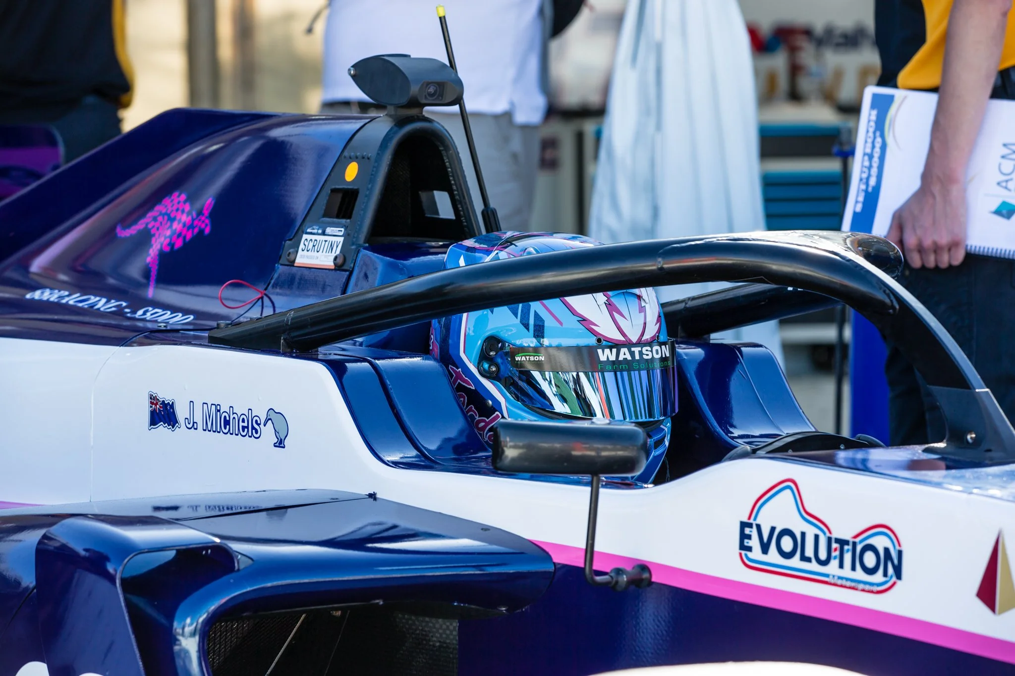 Close-up of a race car with sponsor logos, driver helmet visible inside, and people in the background at a racing event.