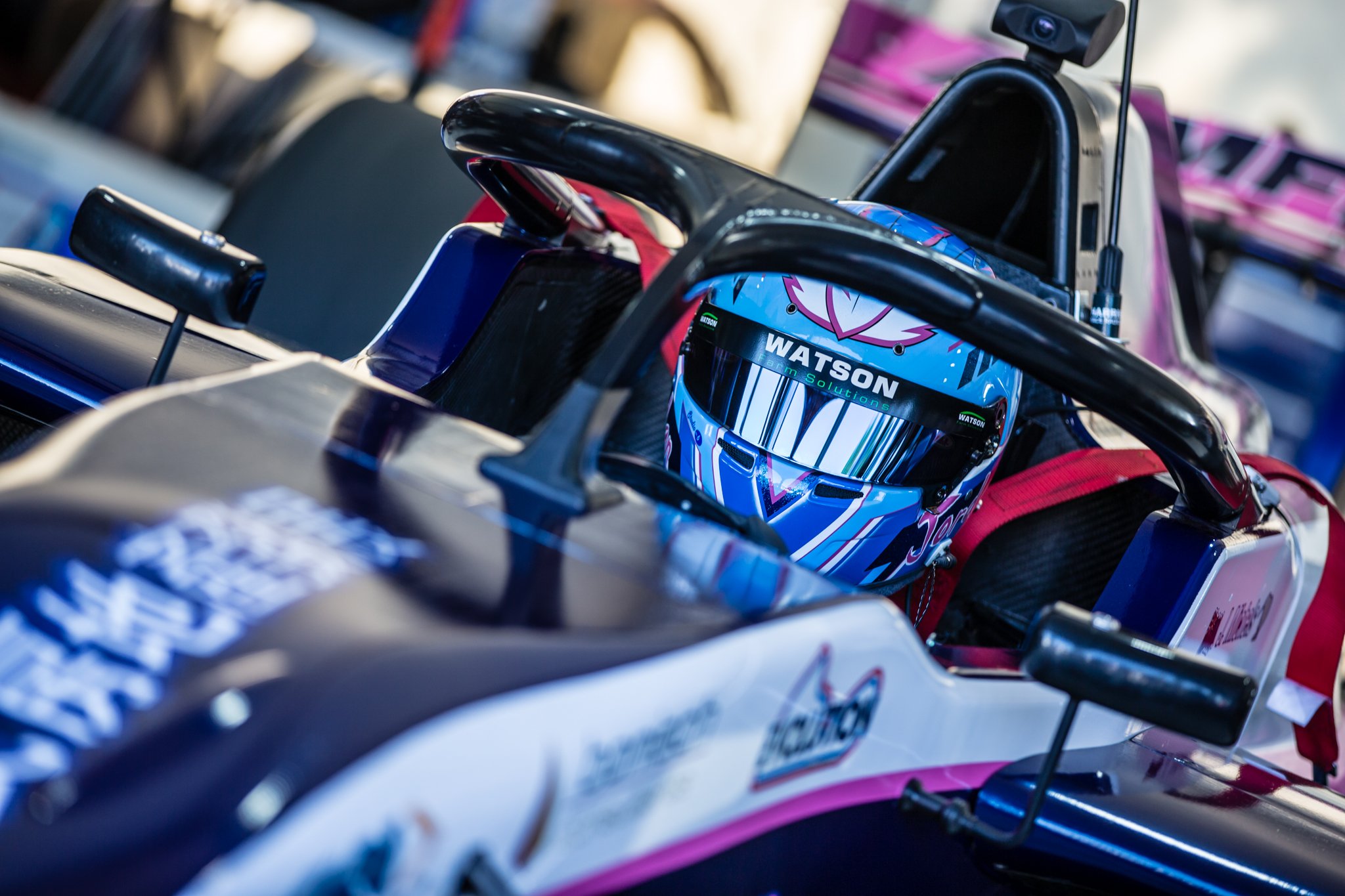 Close-up of a go-kart race car with a blue and pink graphic design, featuring the name Watson on the helmet and parts of the vehicle.