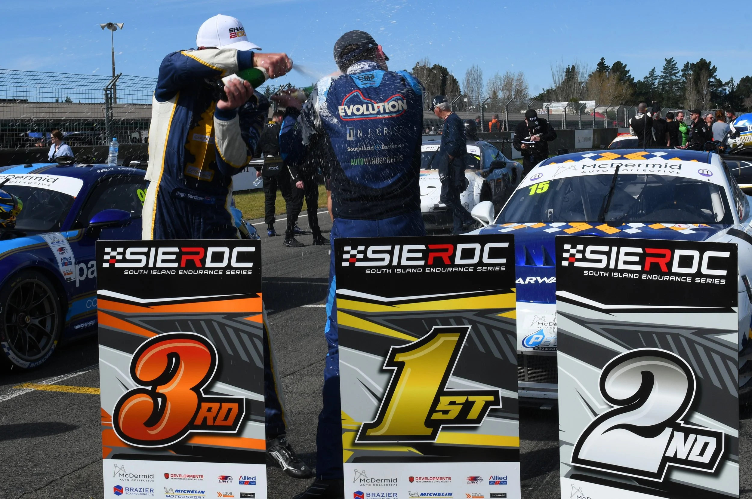 Race car drivers celebrating on the podium, pouring champagne over each other after a race during the South Island Endurance Series, with cars, team members, and race officials present in the background.