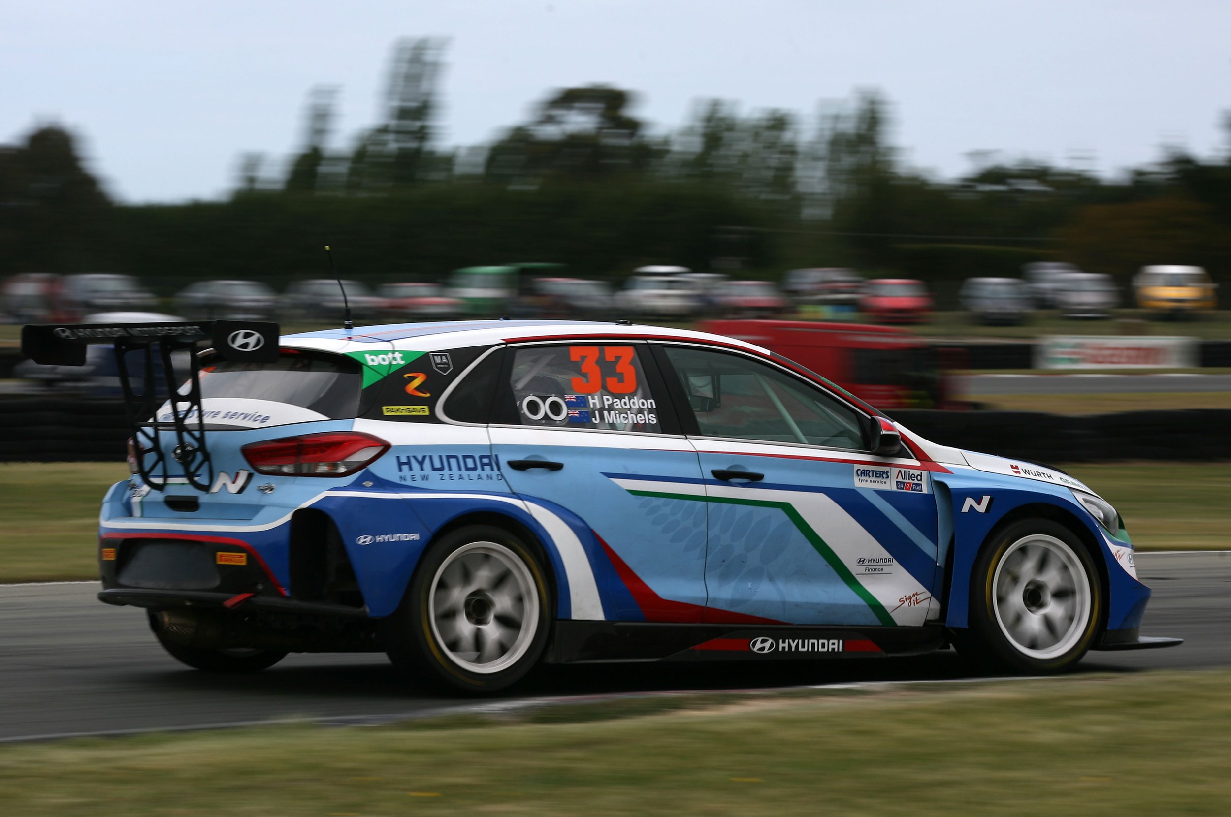 A Hyundai race car with the number 33 is moving quickly on a racetrack, with a blurred background of parked cars and trees.