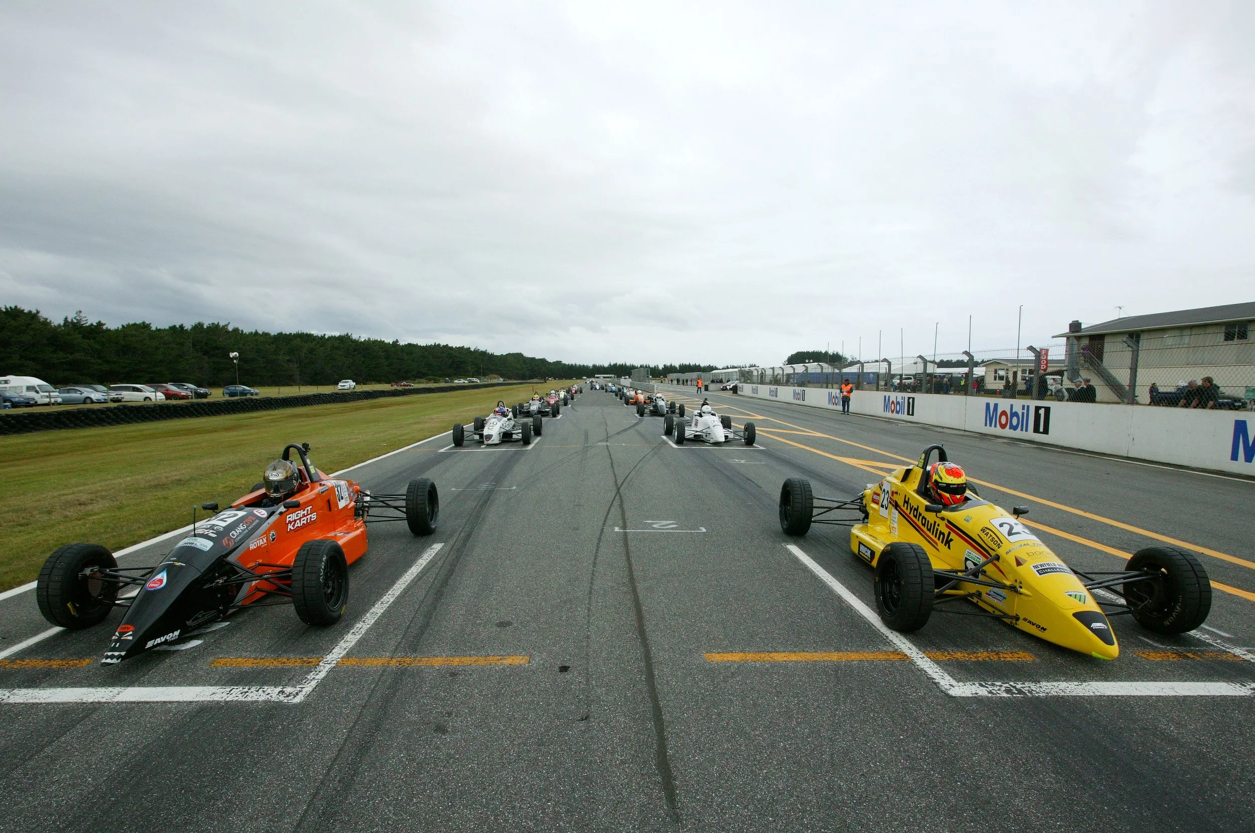 Race cars lined up on the starting grid at a race track, with spectators and officials in the background.