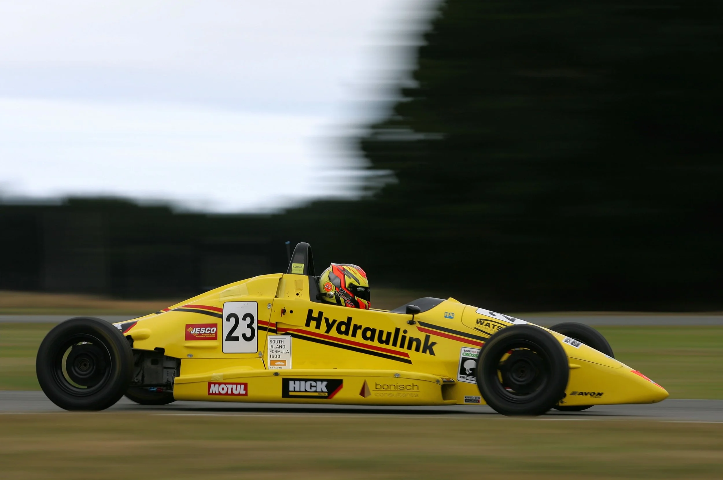 Yellow Formula 1600 race car with racing number 23, sponsored by Hydraulink, moving on a race track with blurred background, driven by a driver wearing a helmet, during a race.