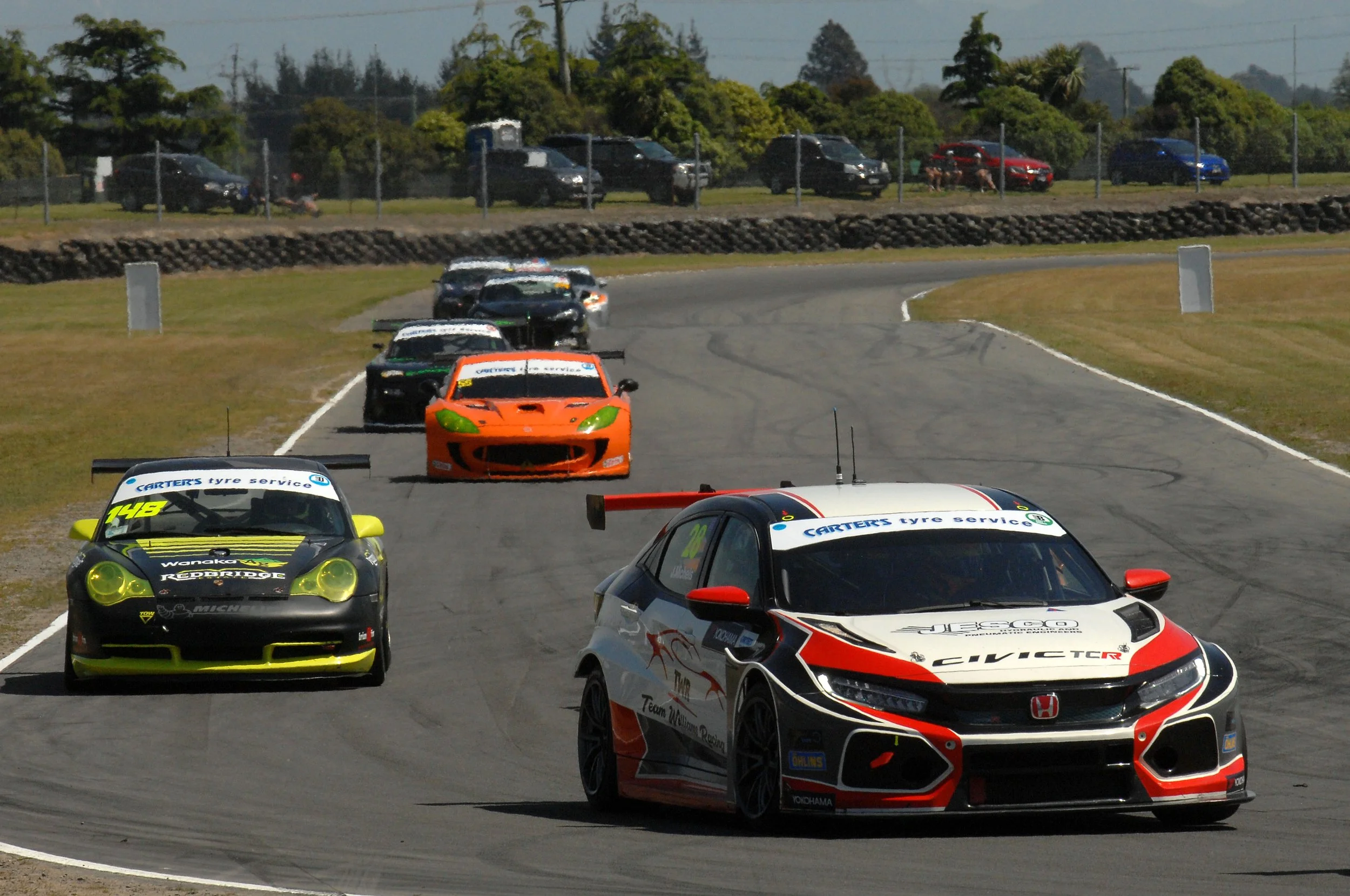 Multiple race cars on a track, with some cars ahead and others following behind, during a race event, with trees and parked cars in the background.