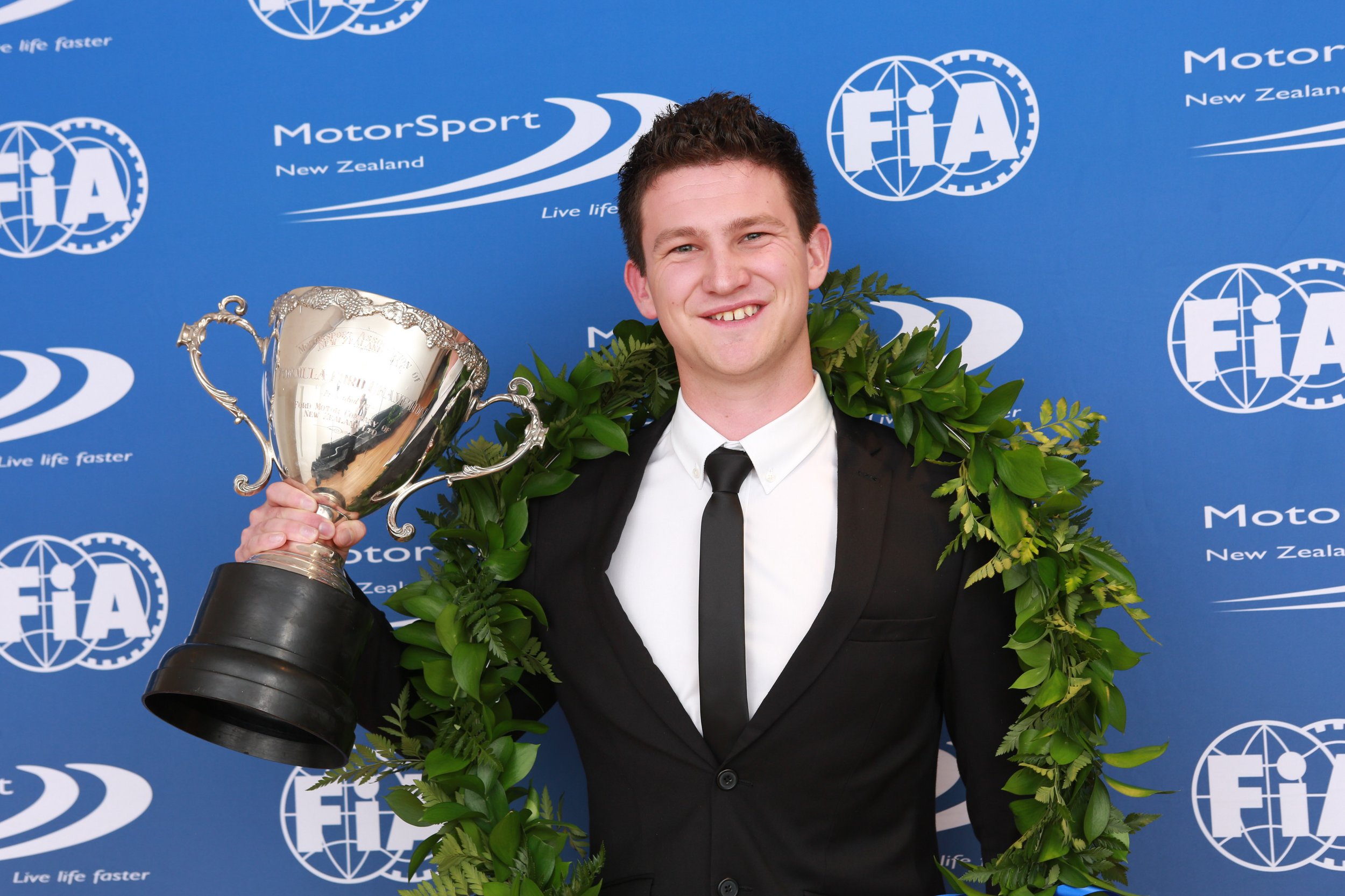 Young man in tuxedo holding a trophy and wearing a laurel wreath, standing in front of a blue backdrop with FIA and MotorSport New Zealand logos.
