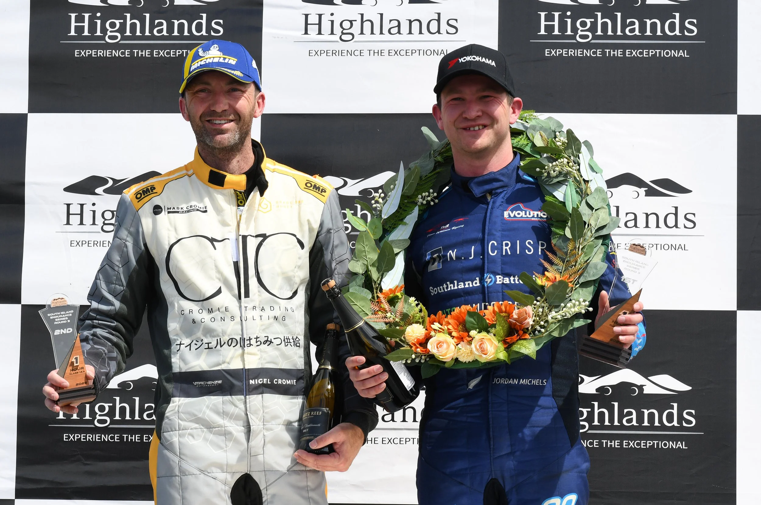 Two race car drivers standing on a podium, smiling, with a black and white checkered backdrop displaying the words "Highlands" and "Experience the Exceptional." The driver on the left is wearing a white racing suit with black and yellow accents, hold