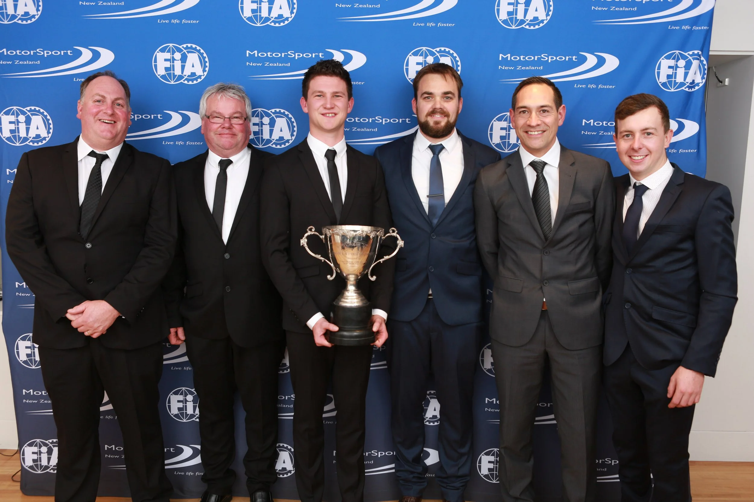 Six men in suits and tuxedos stand in front of a blue backdrop with FIA and MotorSport logos, smiling and posing with a silver trophy.