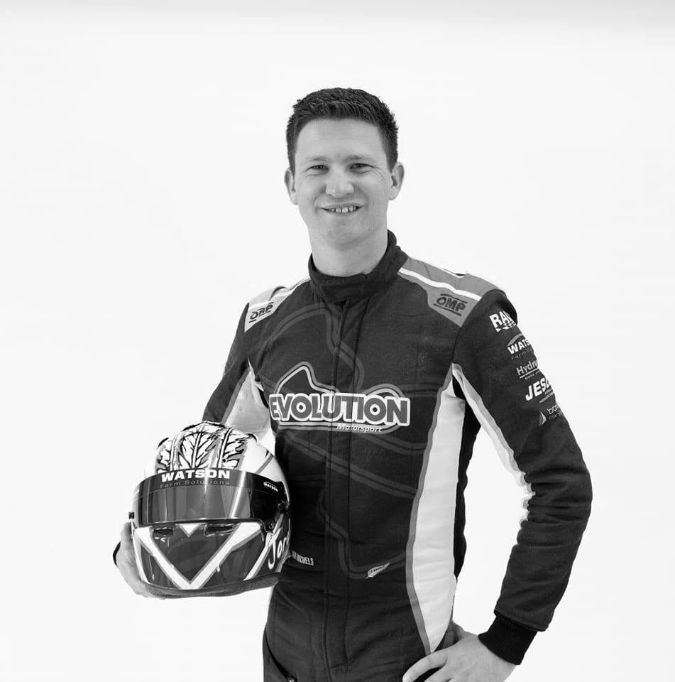 A male race car driver in a racing suit holding a helmet, smiling, in black and white photograph.