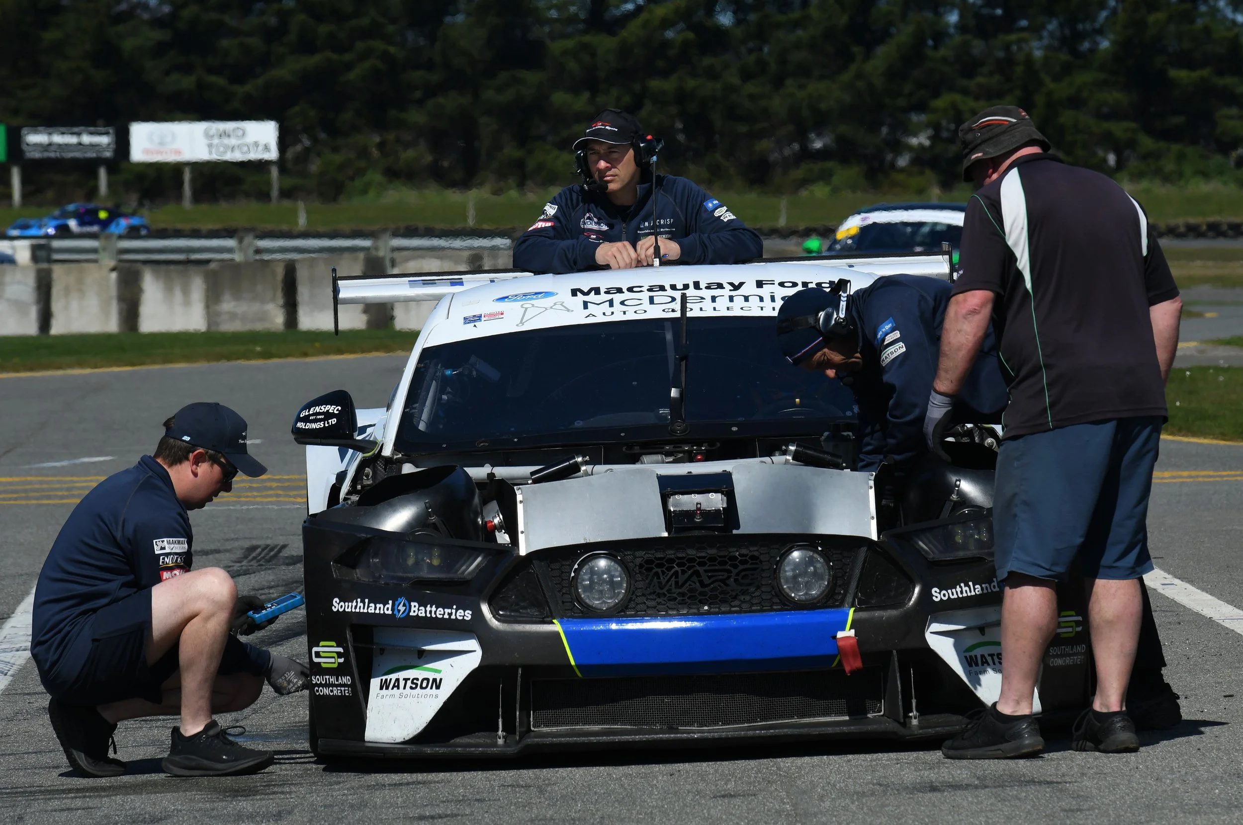 Race car team members working on a white rally car with a large rear wing at a race track, with trees and a blurred race car in the background.