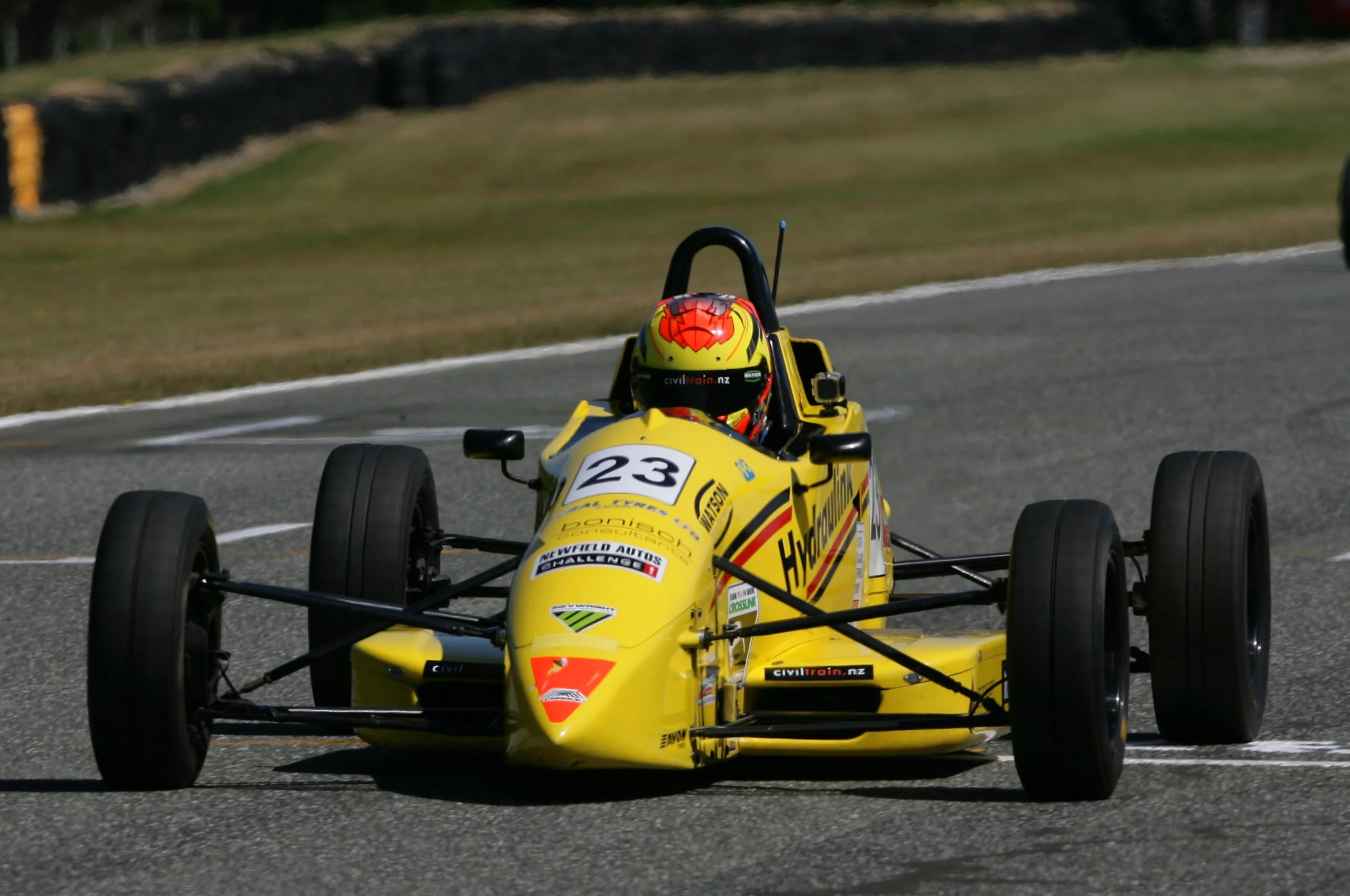 A yellow open-wheel race car on a track, driven by a person wearing a red and yellow helmet, with the number 23 on the front of the car.