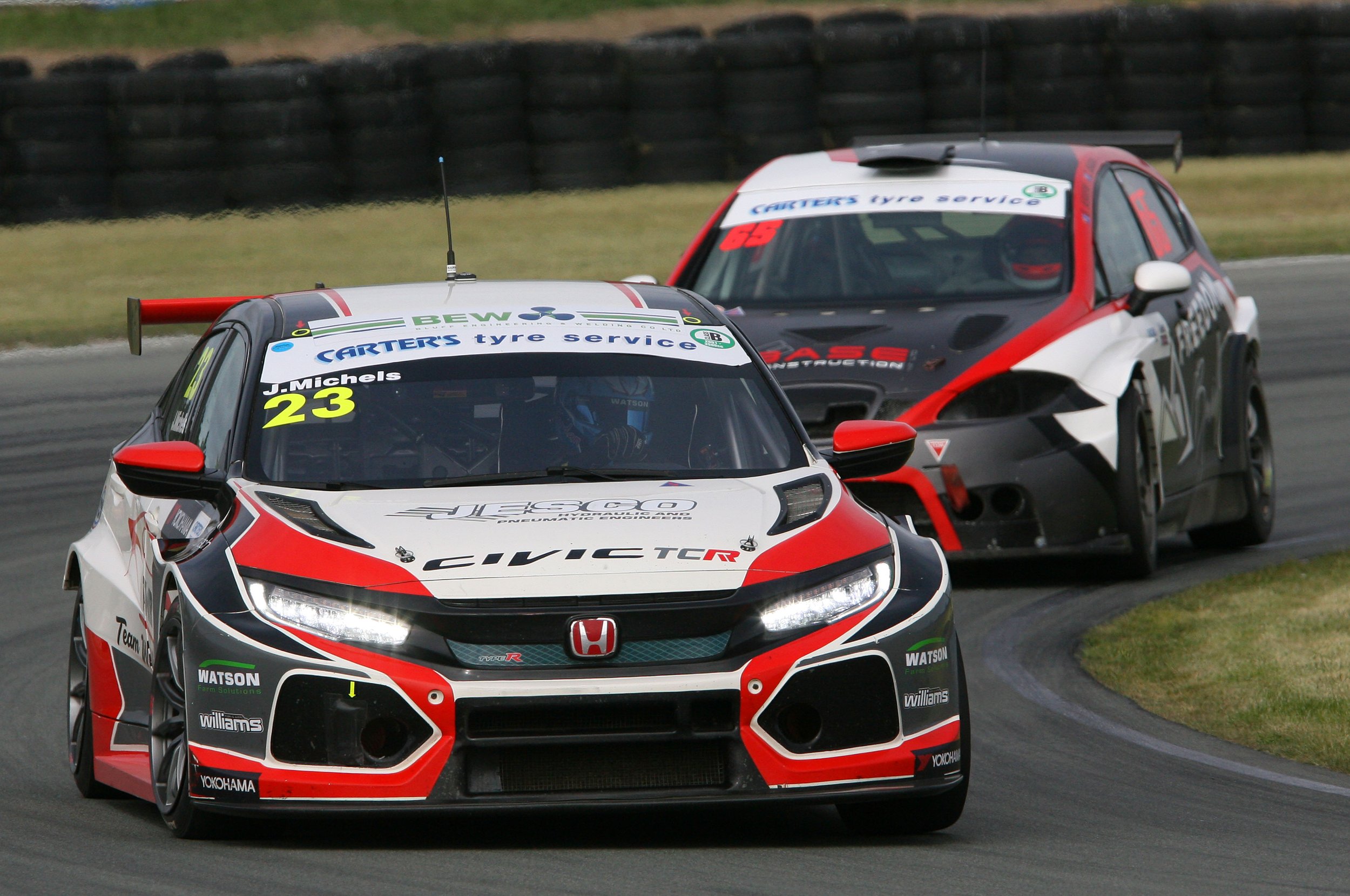 Two race cars on a track, one in front with the number 23, black, white, and red, and the other behind in black, white, and red with a rear wing, racing near a tire barrier.