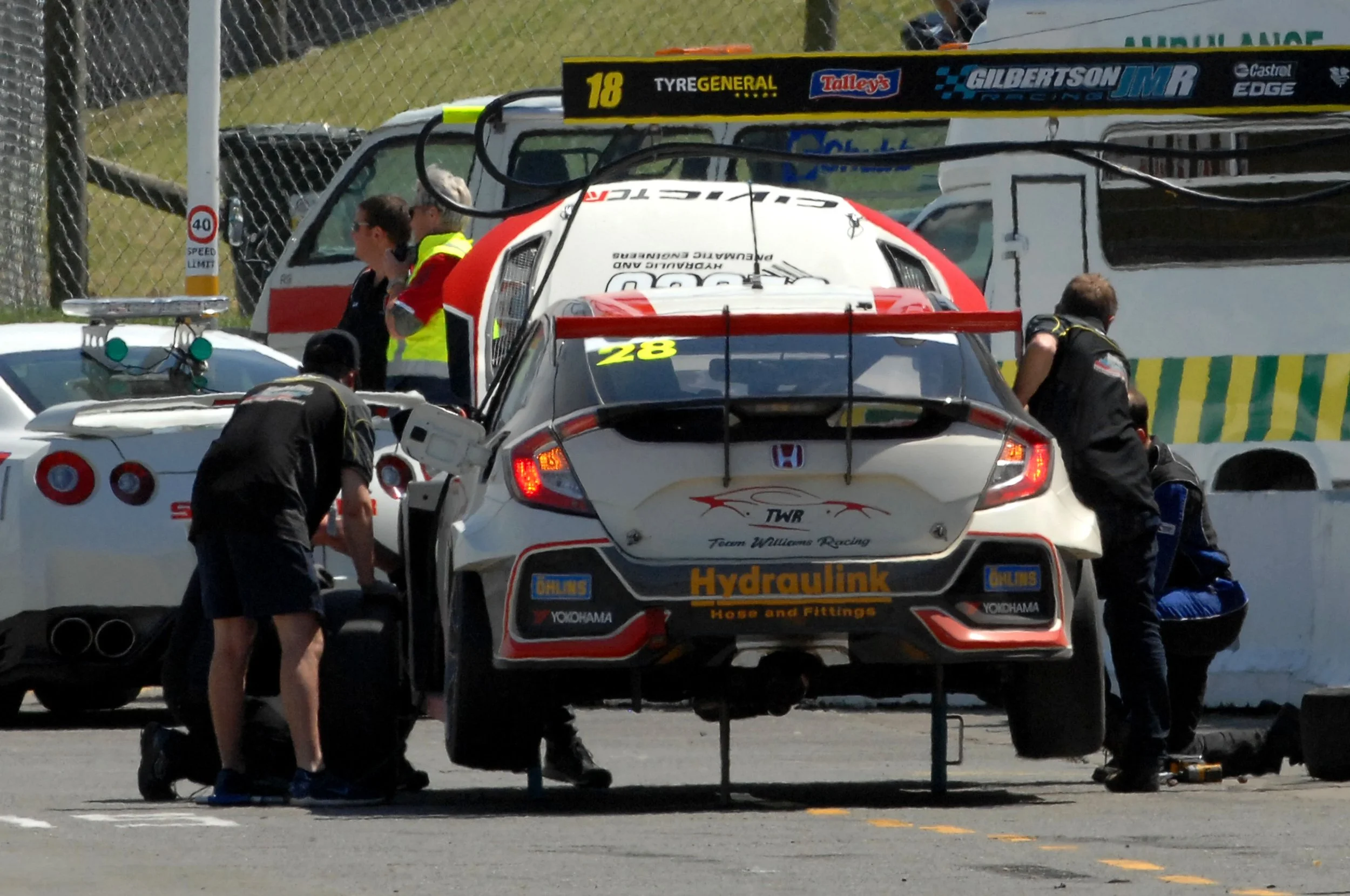 Racecar team members working on car in pit area during a race, with various team and sponsor logos visible.