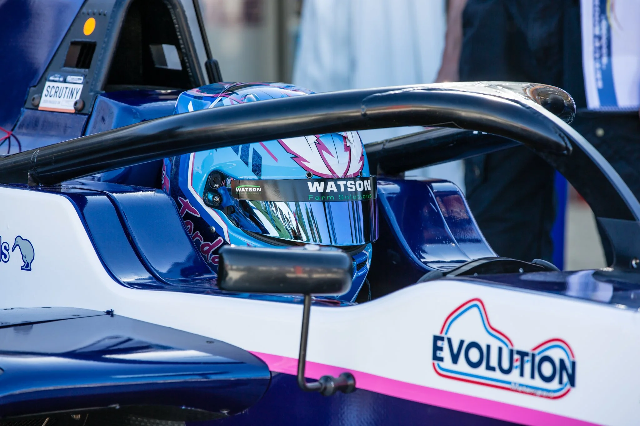 Close-up of a race car driver's helmet inside the cockpit of a race car with sponsor decals, including Watson Farm Solutions, and a portion of a blue and pink livery.