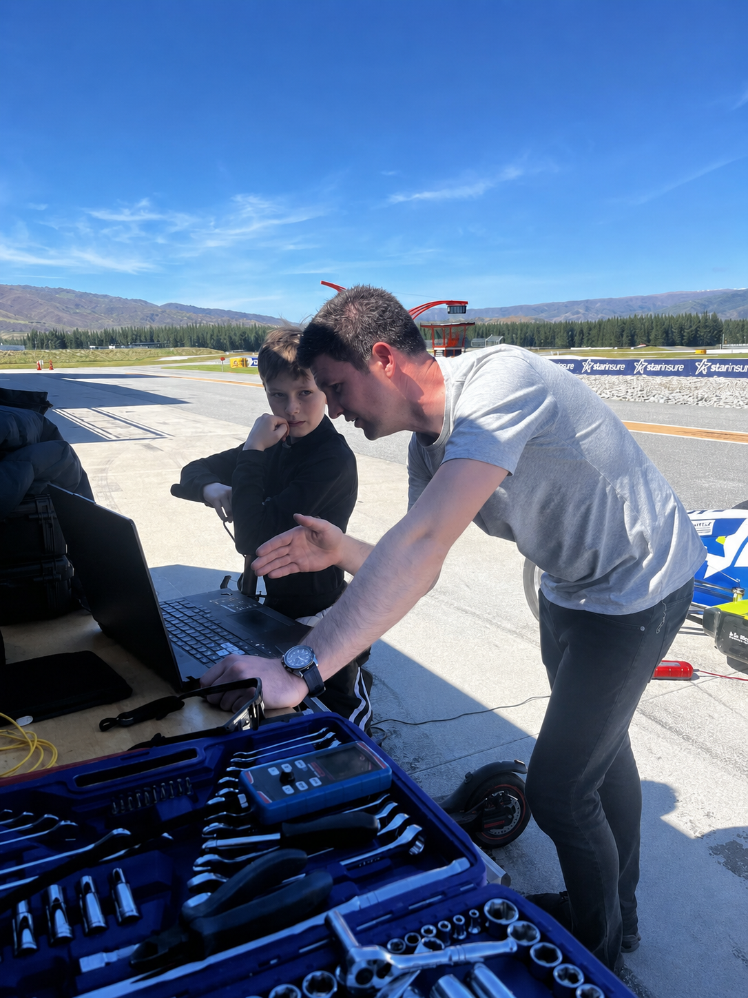 Two men are working together at a motor racing track, with tools and a laptop on a table. One man is explaining something to the other, who appears to be a young boy. The background shows a race track, mountains, and clear skies.