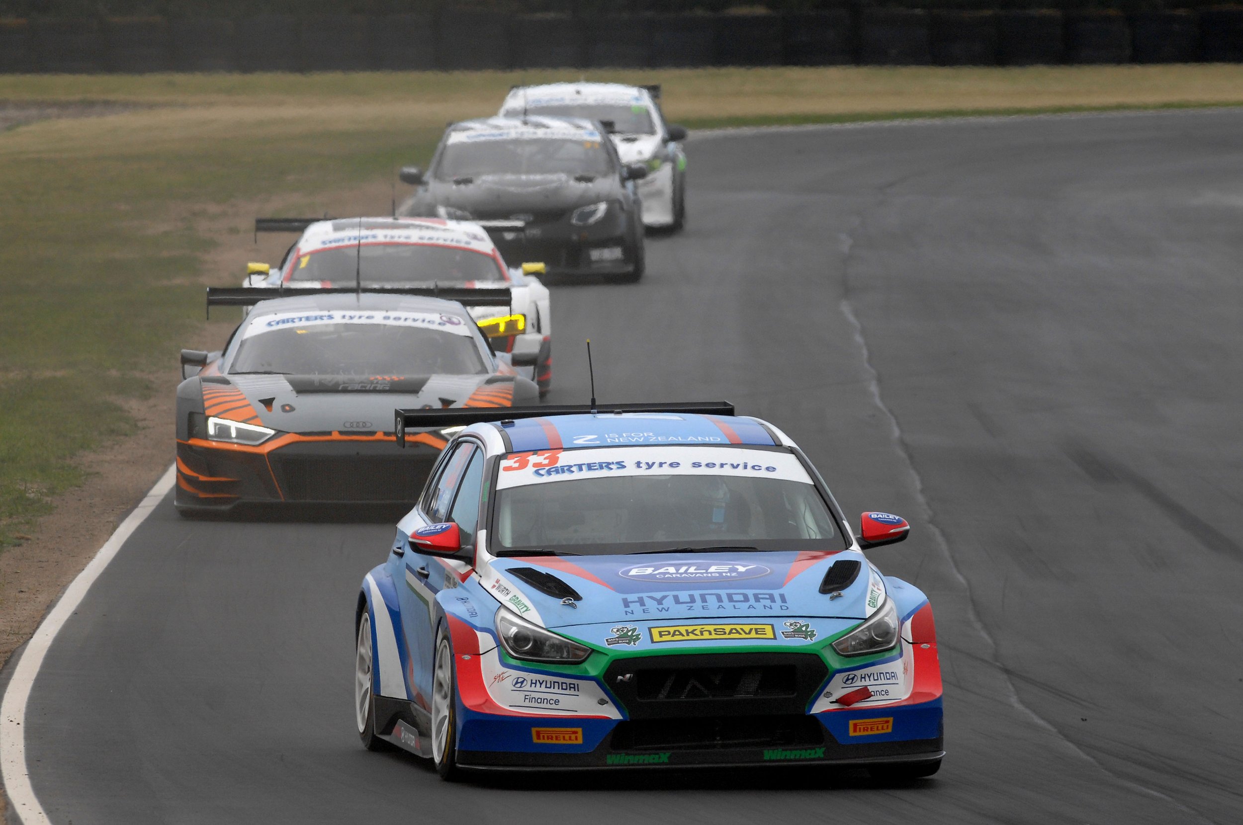 Racing cars in a motorsport race on a paved racetrack, with the front car displaying Hyundai and sponsor logos, followed by other race cars with sponsor branding, all navigating a turn on the track.