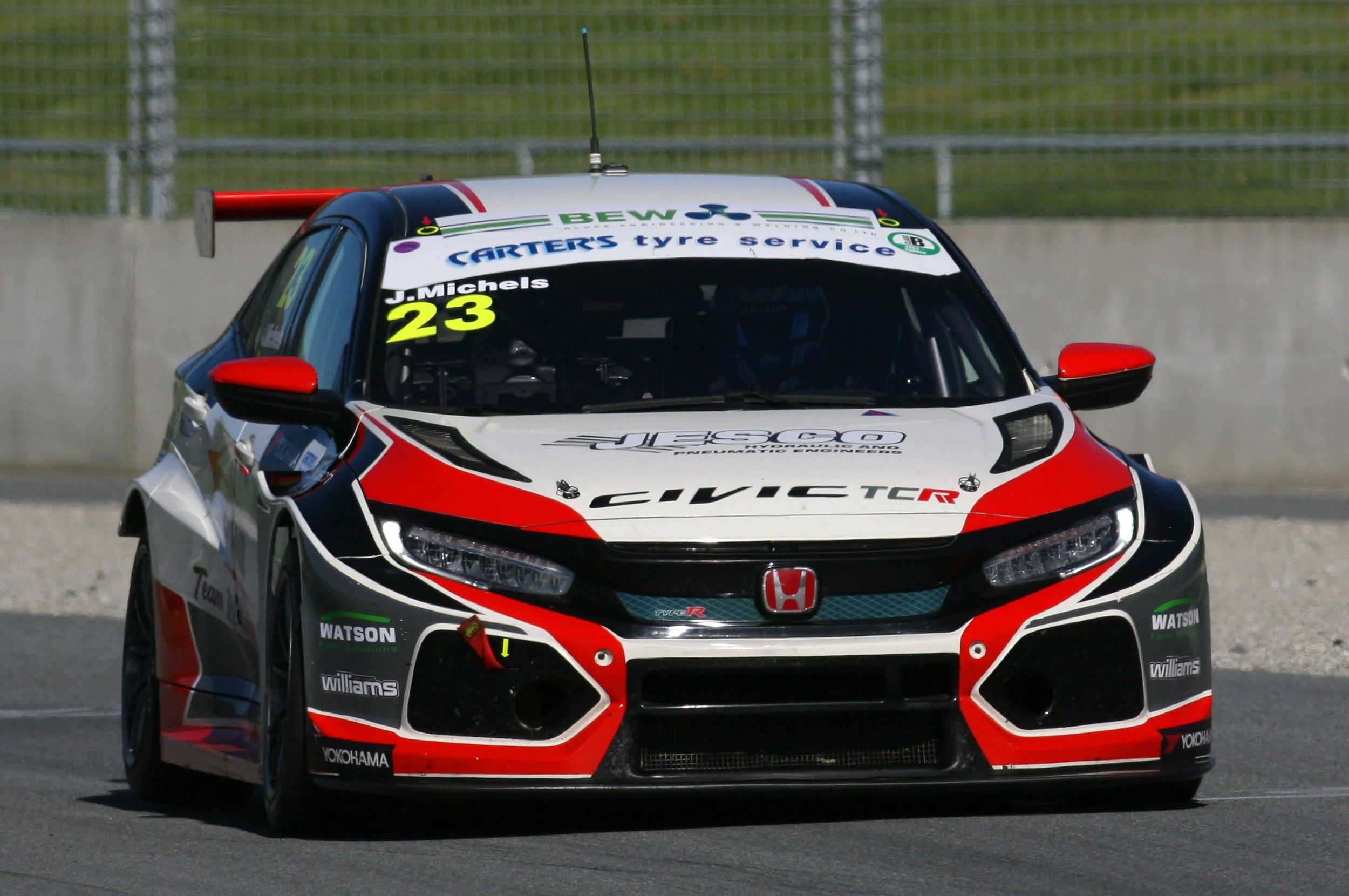 A race car with a red, white, and black design, logo stickers, and the number 23, driving on a track with a green grassy area and a chain-link fence in the background.