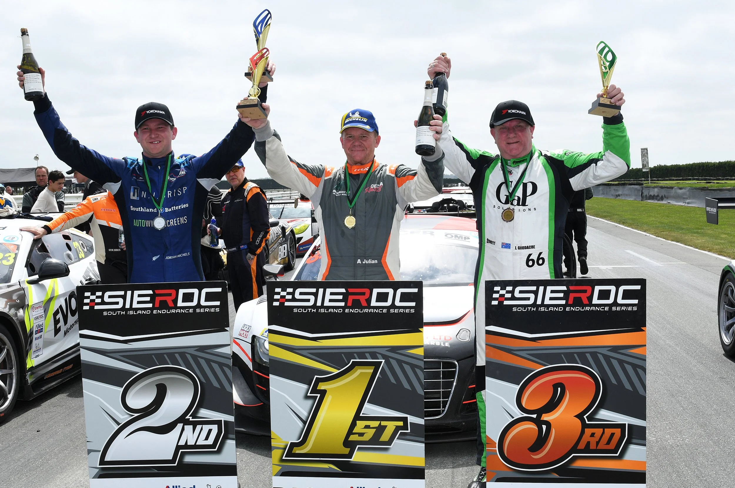 Three race car drivers on a podium celebrating their victory at the South Island Endurance Series, holding trophies and bottles of champagne with race cars and other people in the background.