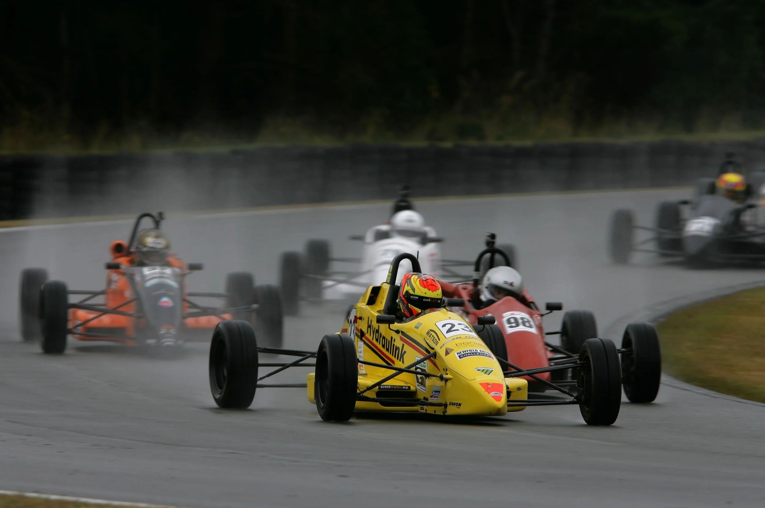 A group of open-wheel race cars on a wet racing track during a race, with the lead car being yellow and others in red, orange, and white, surrounded by greenery and trees.
