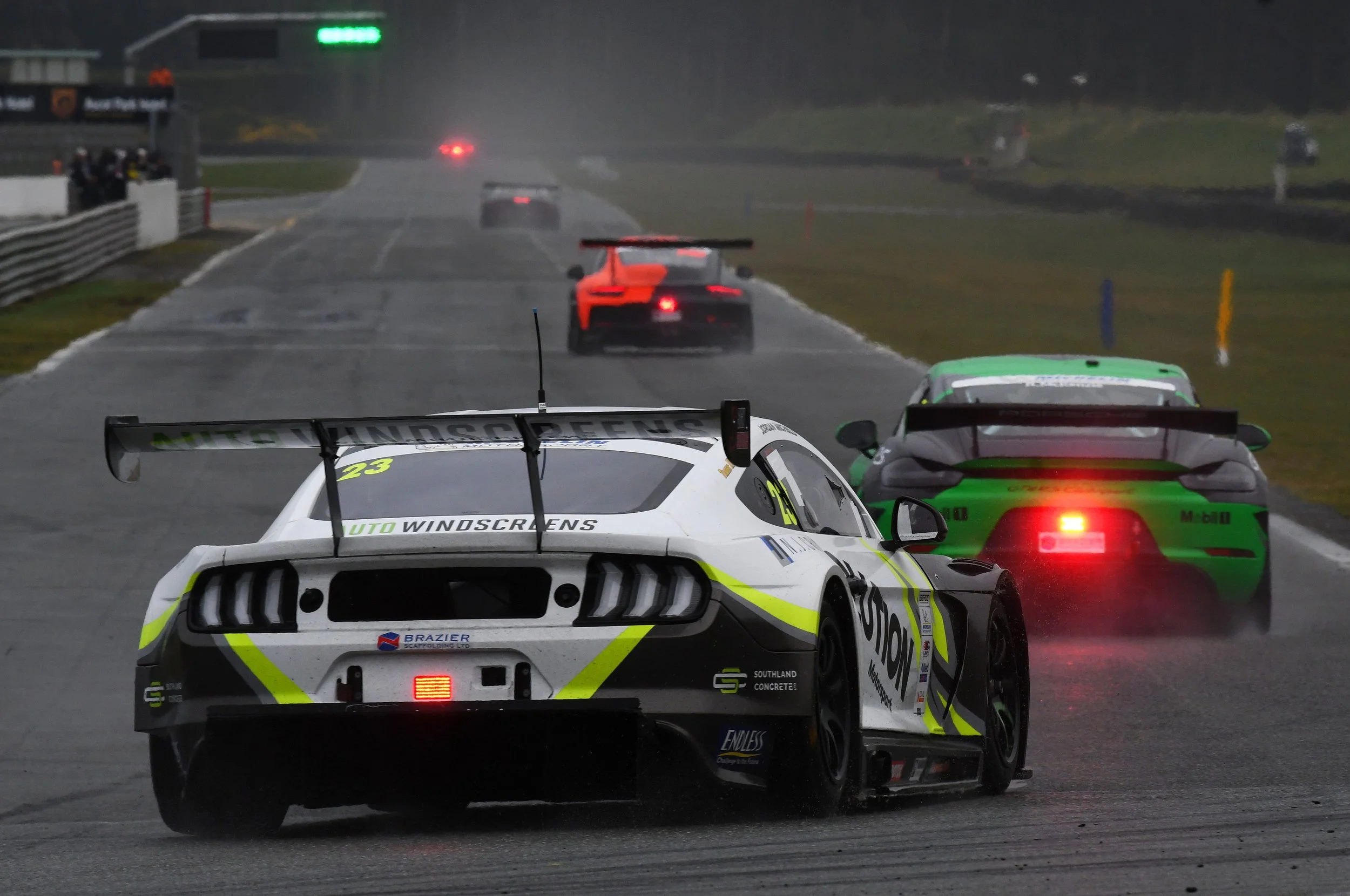 View of race cars on a wet race track during rainy conditions, with blurred background and spectators on the side, showing a racing event.