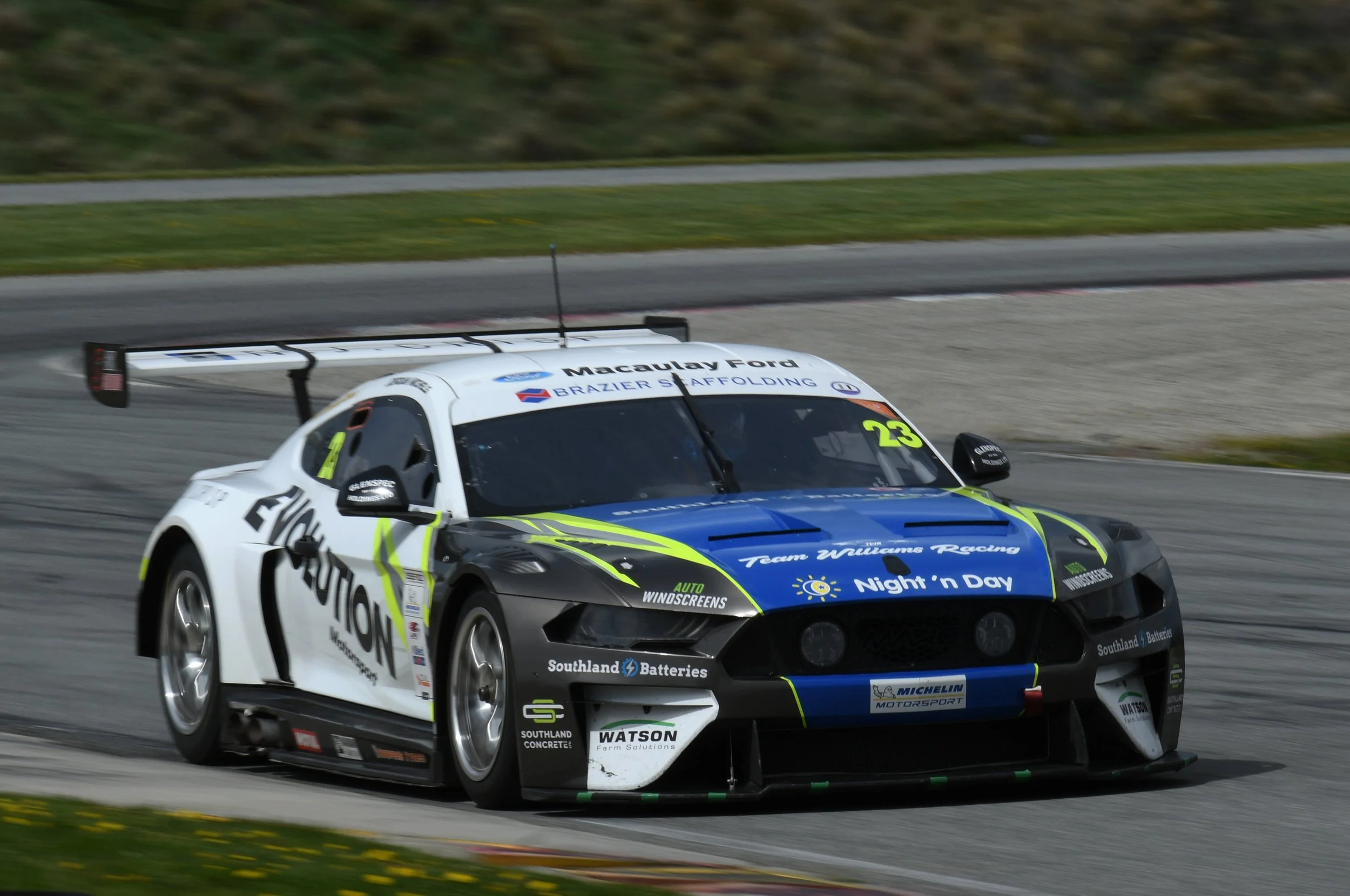 A race car on a track, decorated with various sponsor logos and the number 23, moving at high speed with a blurred background of grass and pavement.