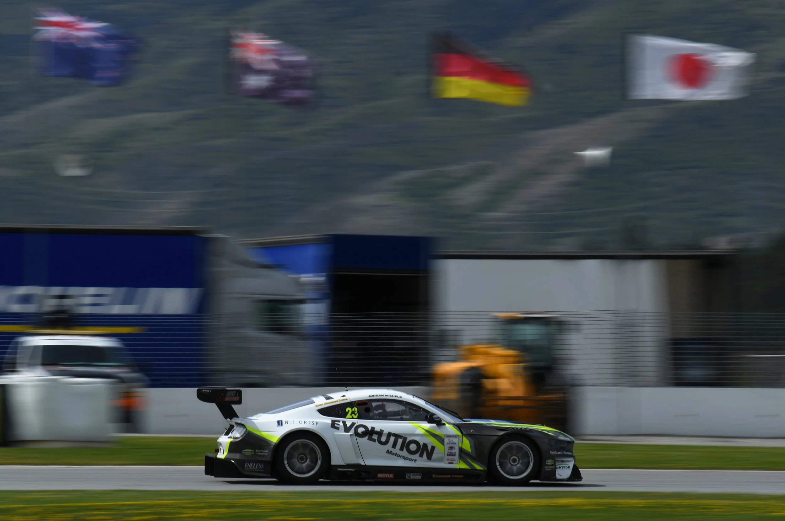 A race car with a white, black, and neon yellow livery, number 23, speeding on a track with a blurred background of flags and machinery.