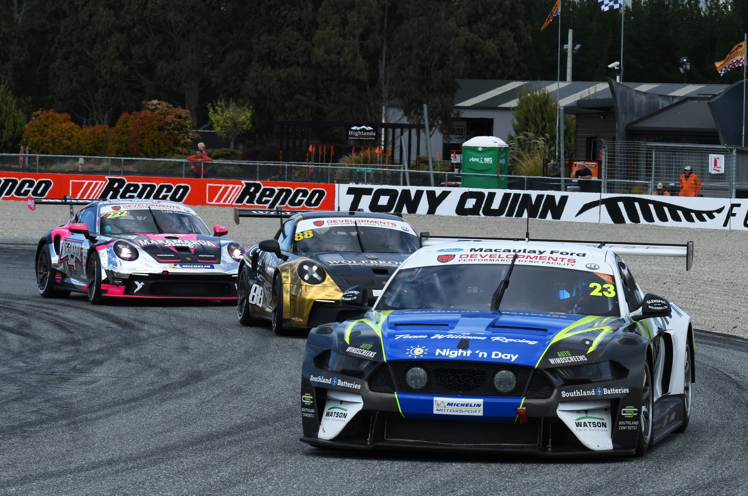 Various race cars on a racetrack, with sponsor banners and spectators in the background.