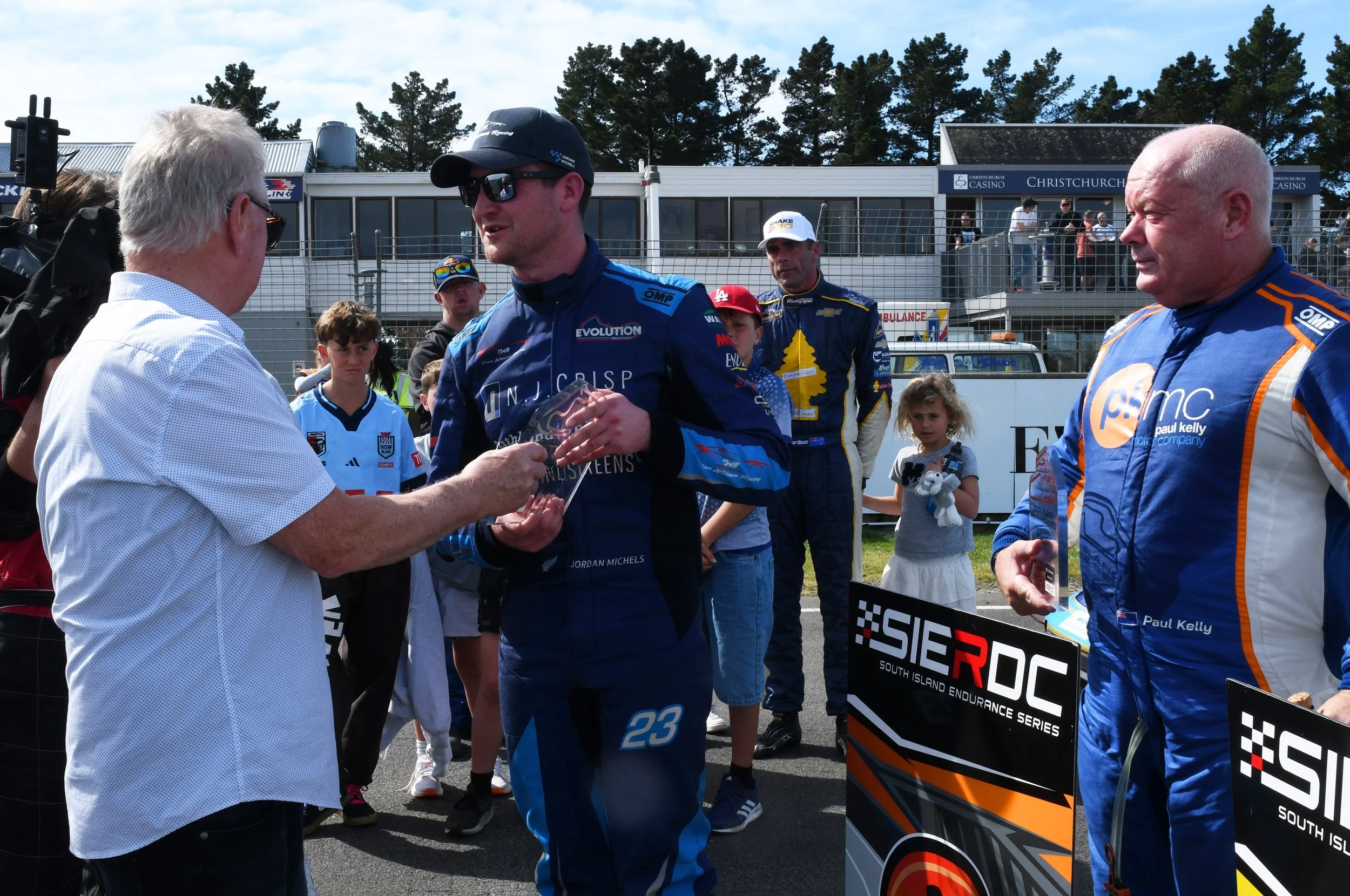 Race car driver in blue racing suit shaking hands with an older man in a white shirt at a racing event, with spectators and team members in the background.