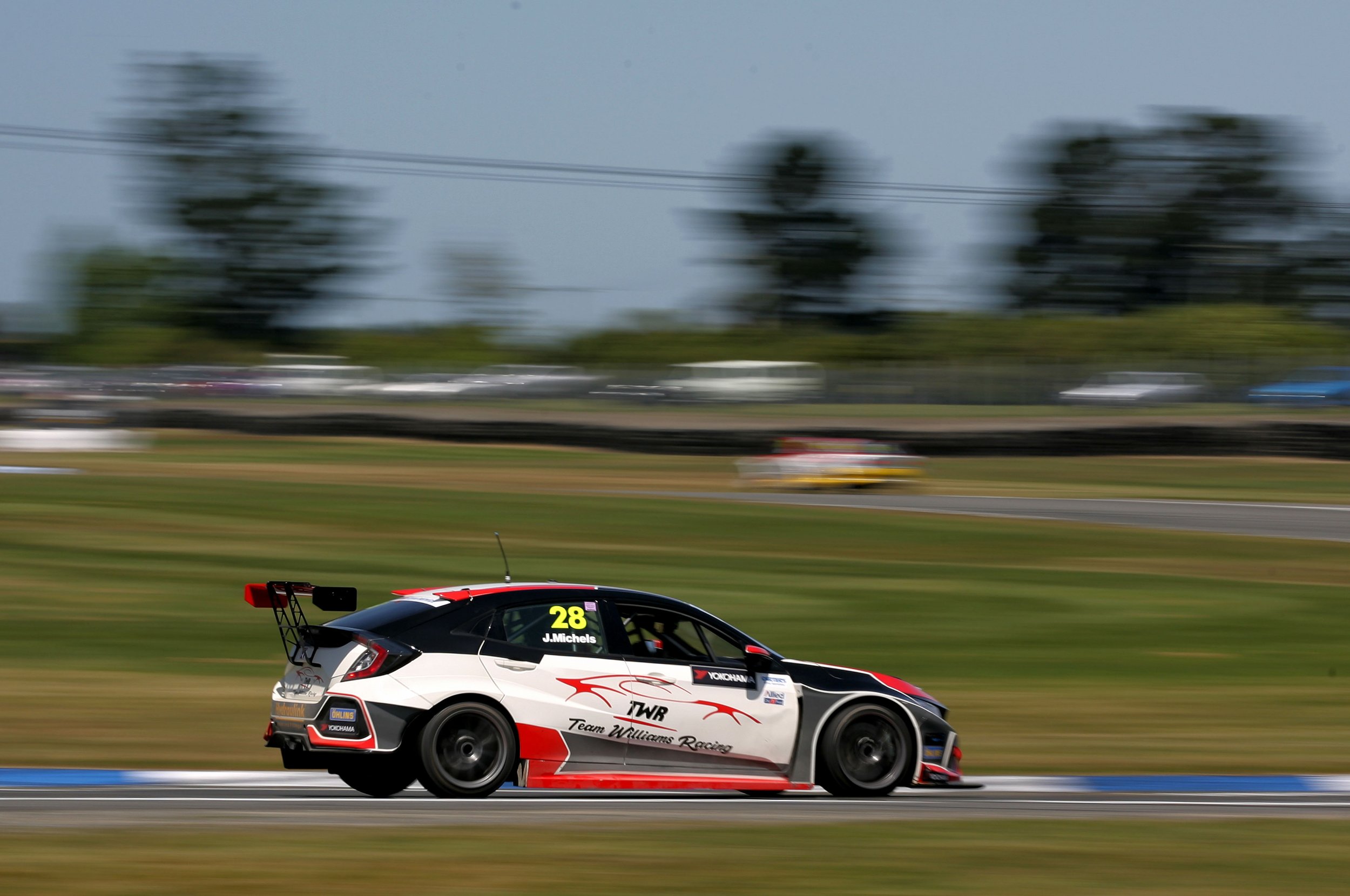A red, white, and black race car with the number 28 on the side, driven by J. Michels, speeding on a race track during a race in daytime, with a blurry background of trees and spectators.