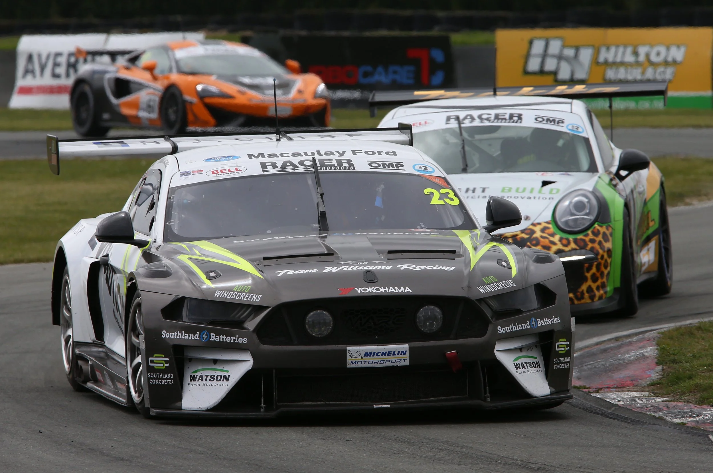 Race cars on a track, with the closest being a gray sports car with various sponsor logos and a large rear wing, followed by an orange and black race car, and a white race car with rainbow and leopard print accents in the background.