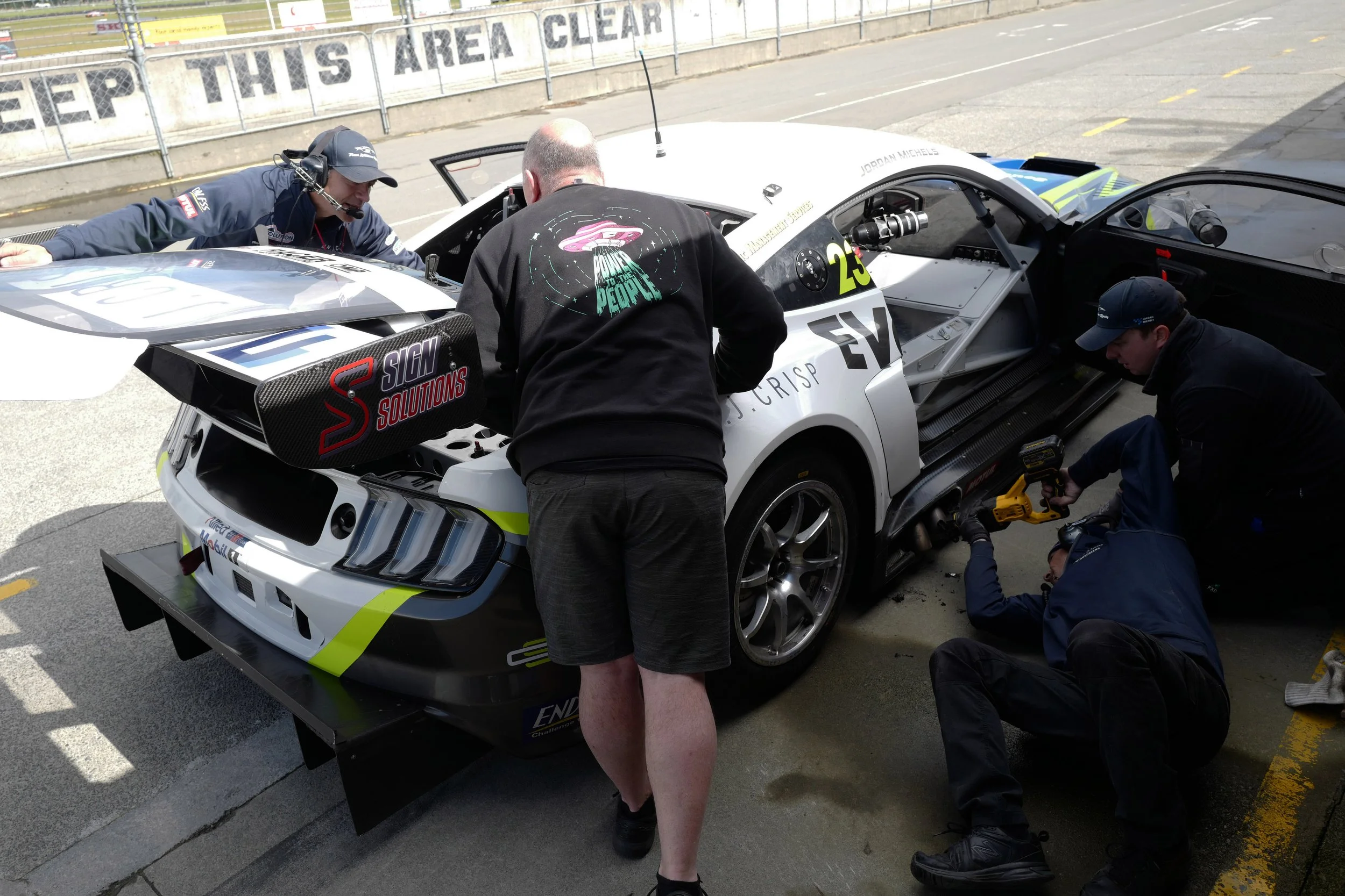 A race car in a pit stop with three crew members working on it; one is wearing a headset and cap, another with a black jacket with colorful graphics on the back, and the third is crouched with a wrench, all focused on the car's front.
