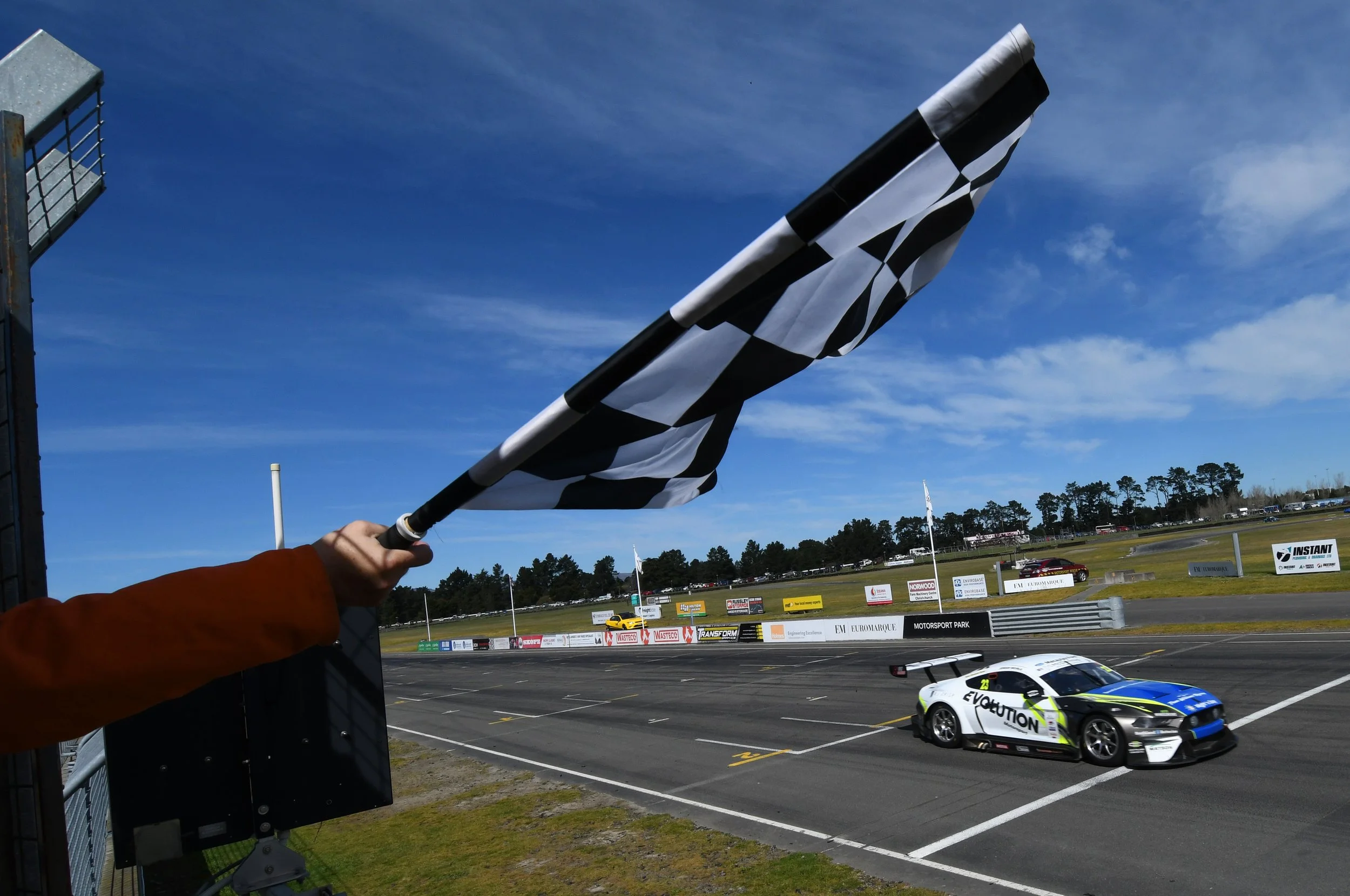 A person holding a black and white checkered racing flag at a motorsports event. Several race cars are visible on the track, including a white and green car with the word 'Evolution' on it. The background features billboards, a clear blue sky, and trees.