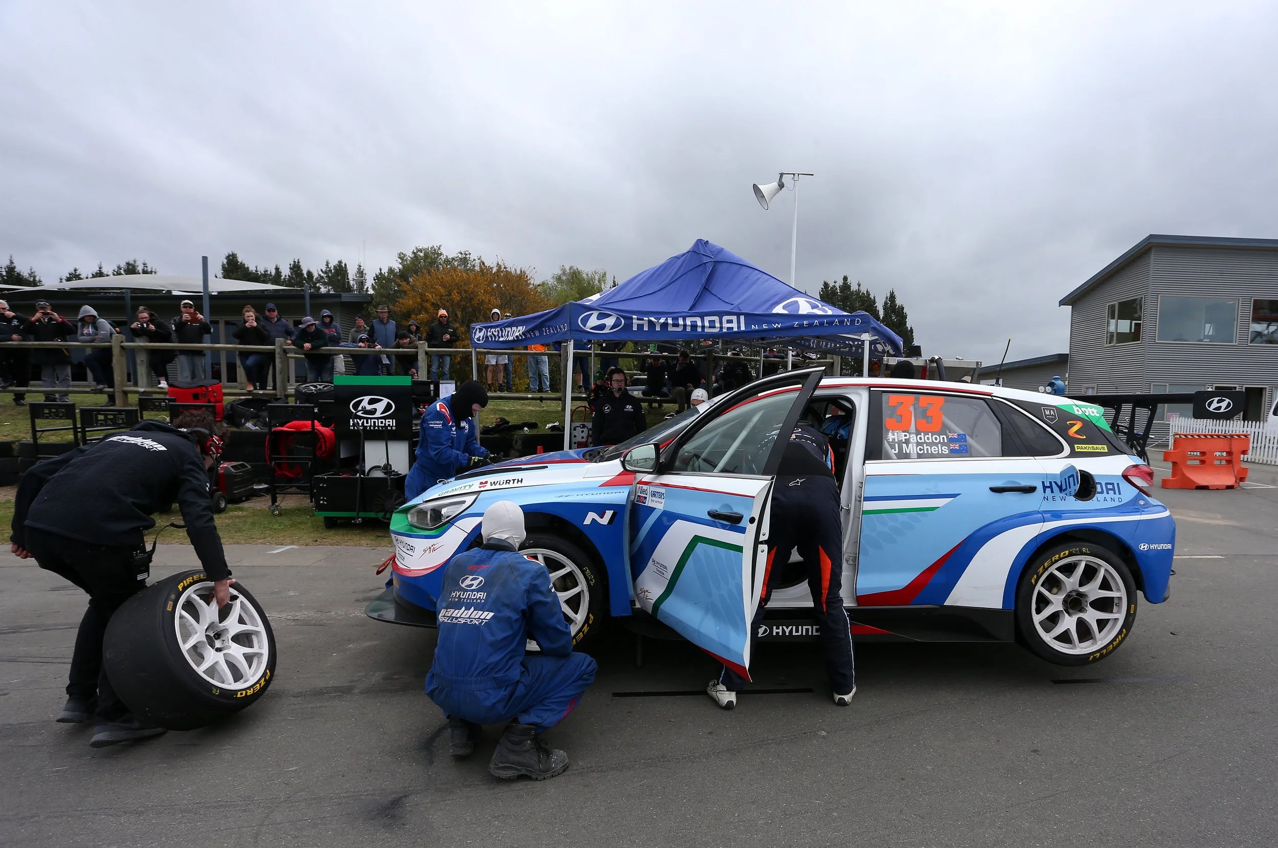 A race car is being repaired in a pit stop area with mechanics working on it. The car is blue with green, red, and white accents and the number 33 on the window. There are people watching from a fenced area, under a blue tent with Hyundai branding, o