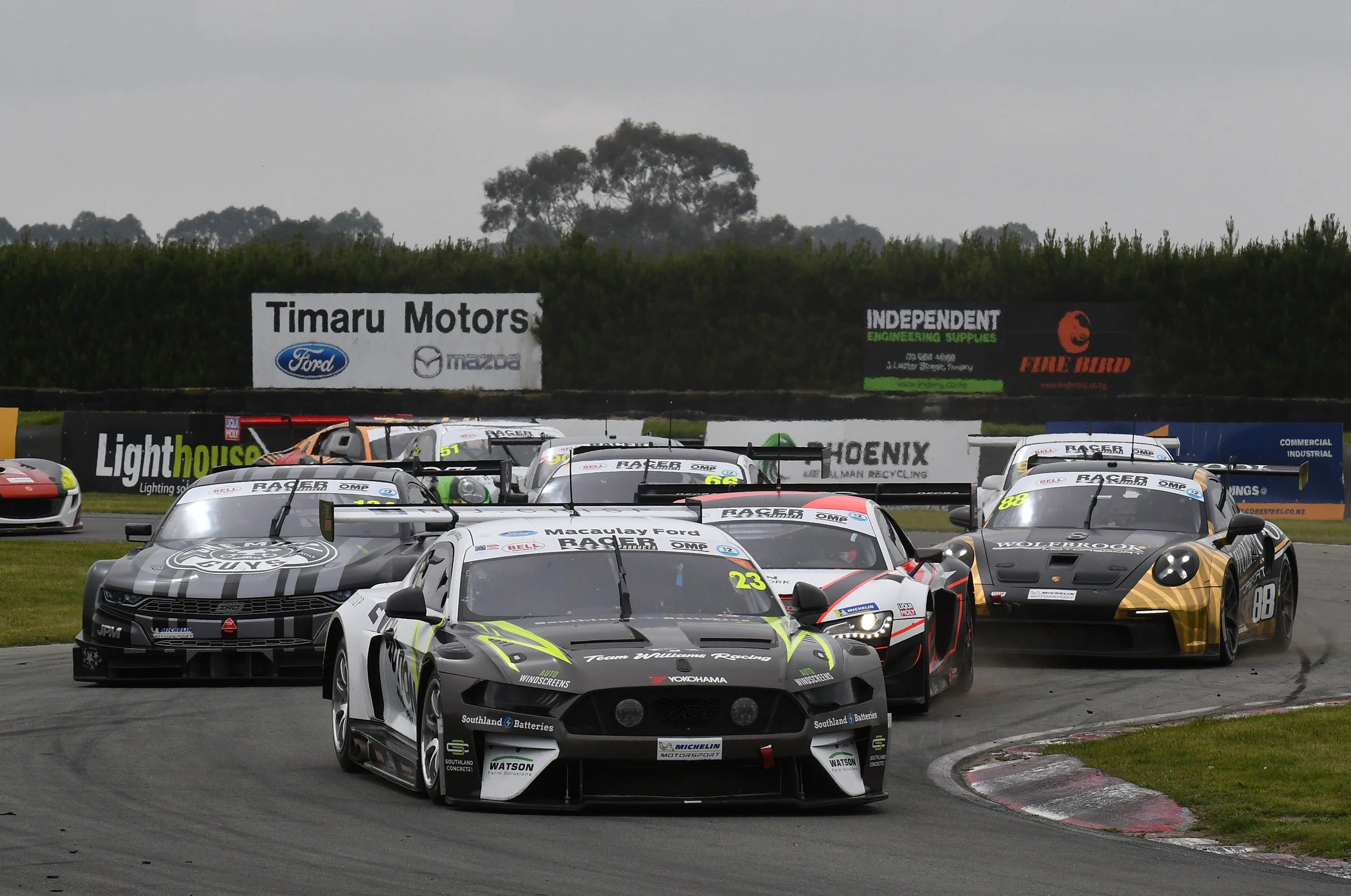 Multiple race cars on a track during a race, with signage and trees in the background.