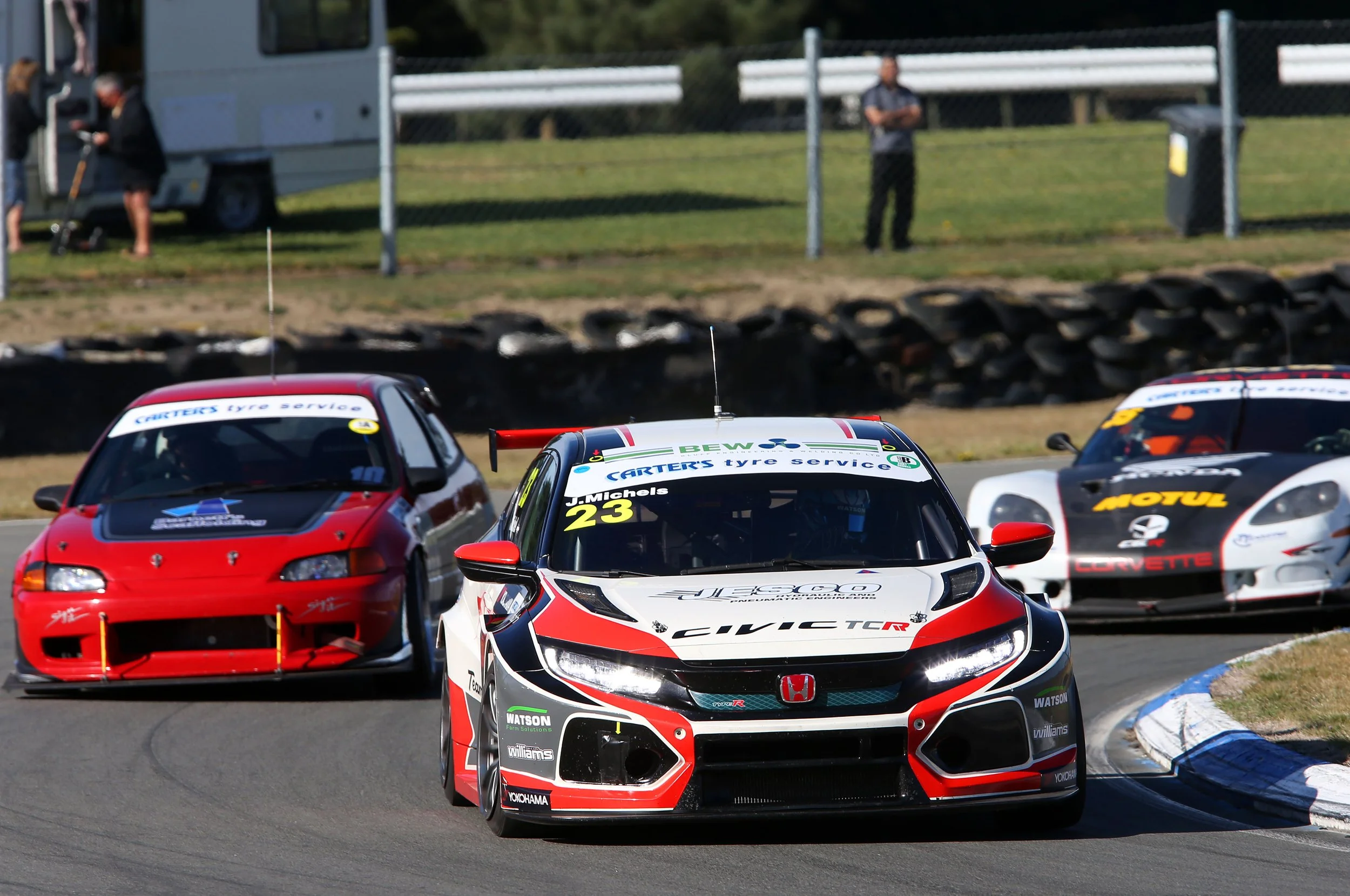 Race cars on a track, including a white and red Honda Civic Type R and a red car, navigating a corner with tire barriers and a green grassy area in the background, along with some spectators.