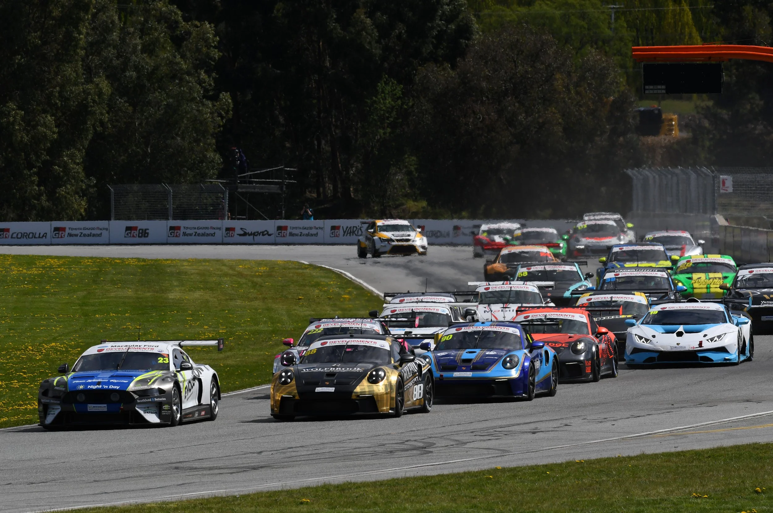 Multiple race cars in various colors on a racing track during a motorsport event, with trees and safety barriers in the background.