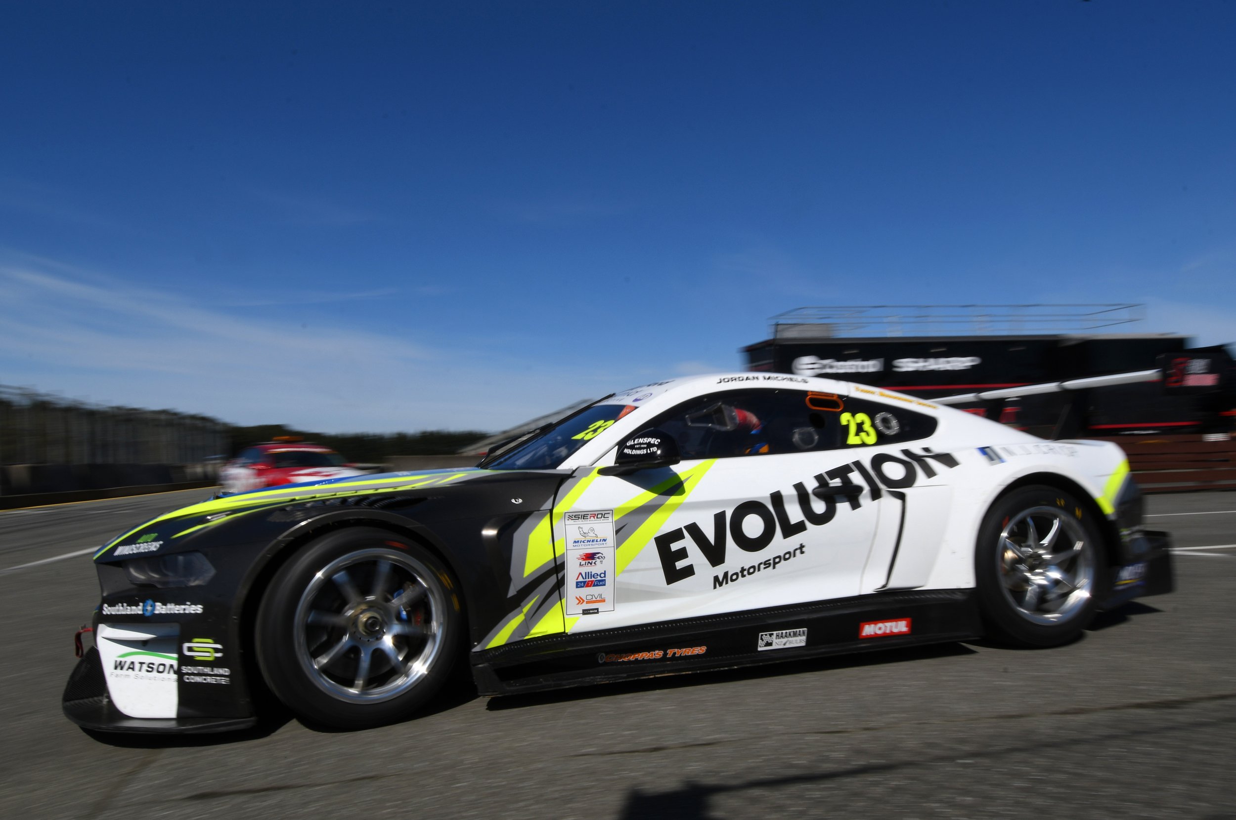 A racing car with a white, black, and yellow livery speeding on a race track under a clear blue sky.
