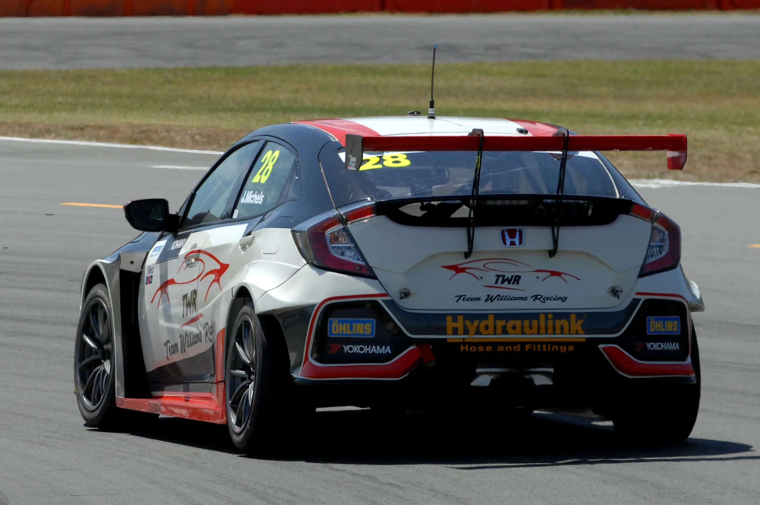 A race car on a track with the number 28 and the name J. Michels on the side, featuring a prominent red rear wing, sponsor logos, and a white, black, and red color scheme.
