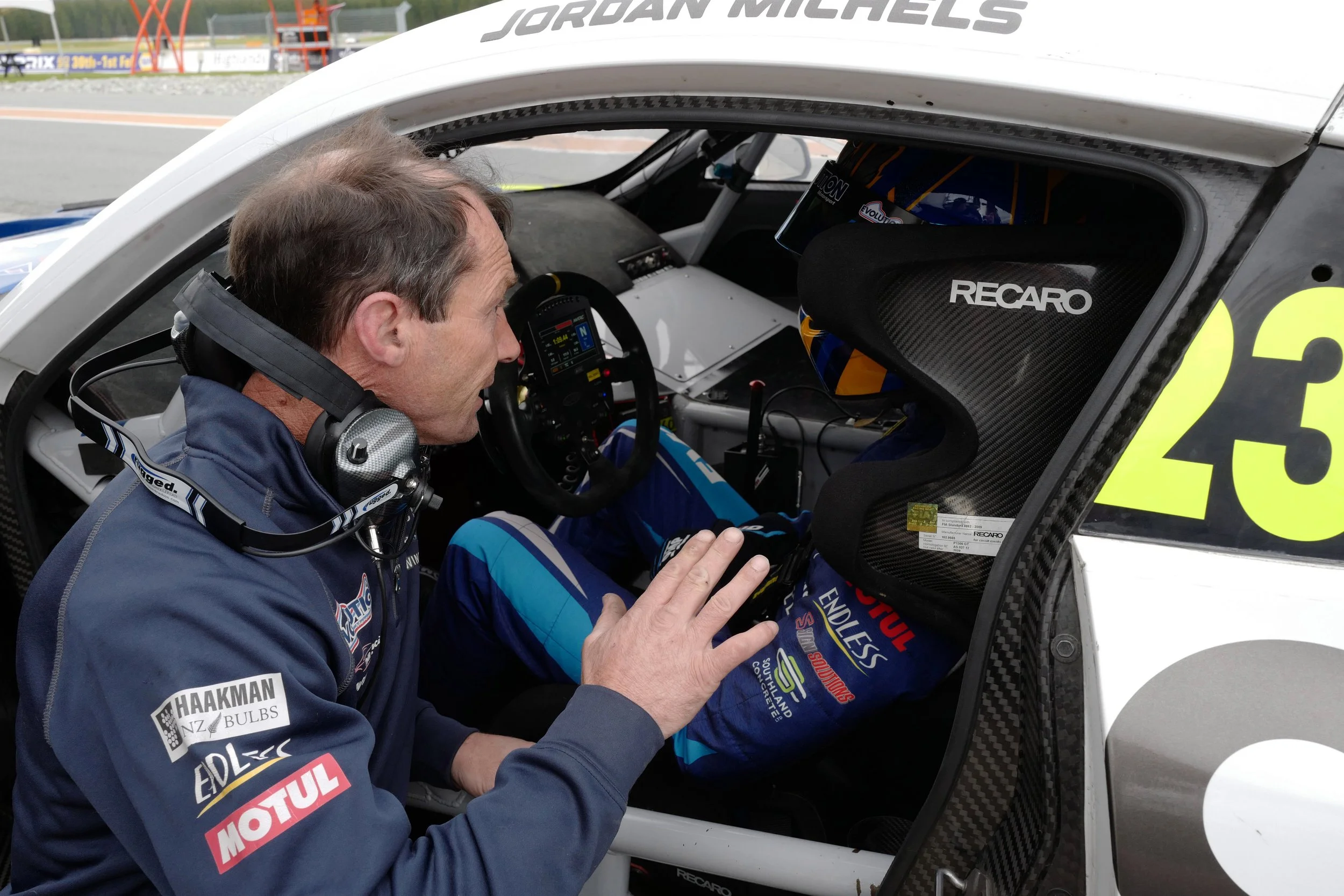 A race car driver, wearing a racing suit with sponsor patches, sits inside a race car with a helmet on, being assisted by a crew member wearing a racing jacket, at a racetrack.