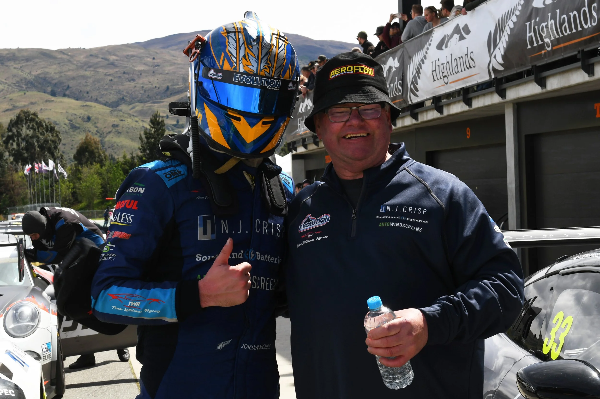 A race car driver in a blue racing suit and helmet giving a thumbs-up, standing next to a man with glasses and a hat, holding a water bottle, at a racing event with spectators and mountains in the background.