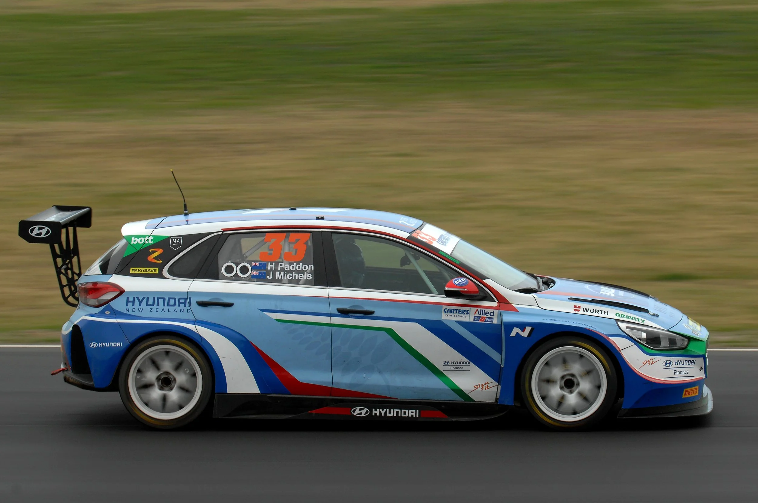 A Hyundai racing car with number 33 and sponsorship logos, speeding on a track with a green grassy background.