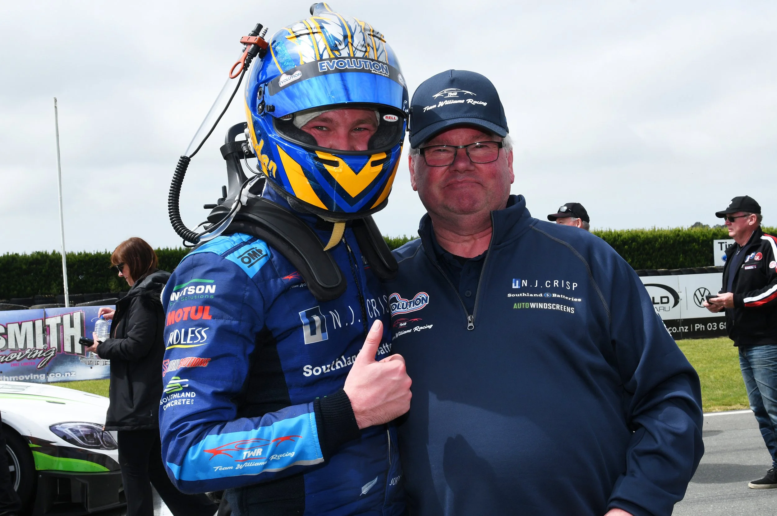 A race car driver in a blue racing suit and helmet posing with a man in a navy blue jacket at a racing event outdoors on a cloudy day.