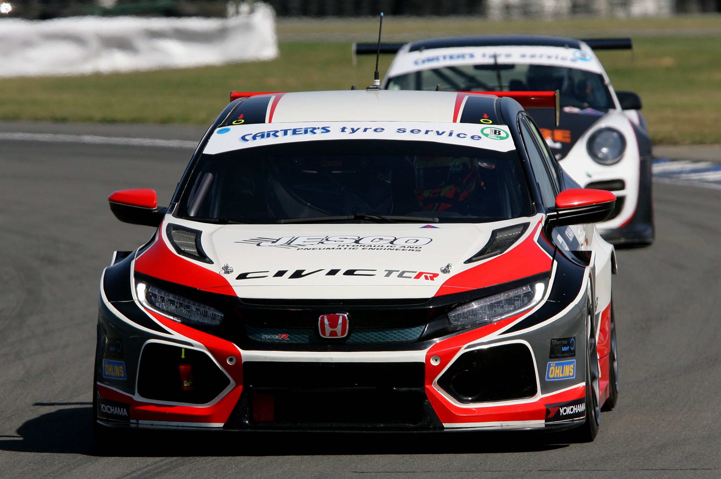 Front view of a red, white, and black Honda Civic Type R race car on a racetrack, with a white race car in the background.