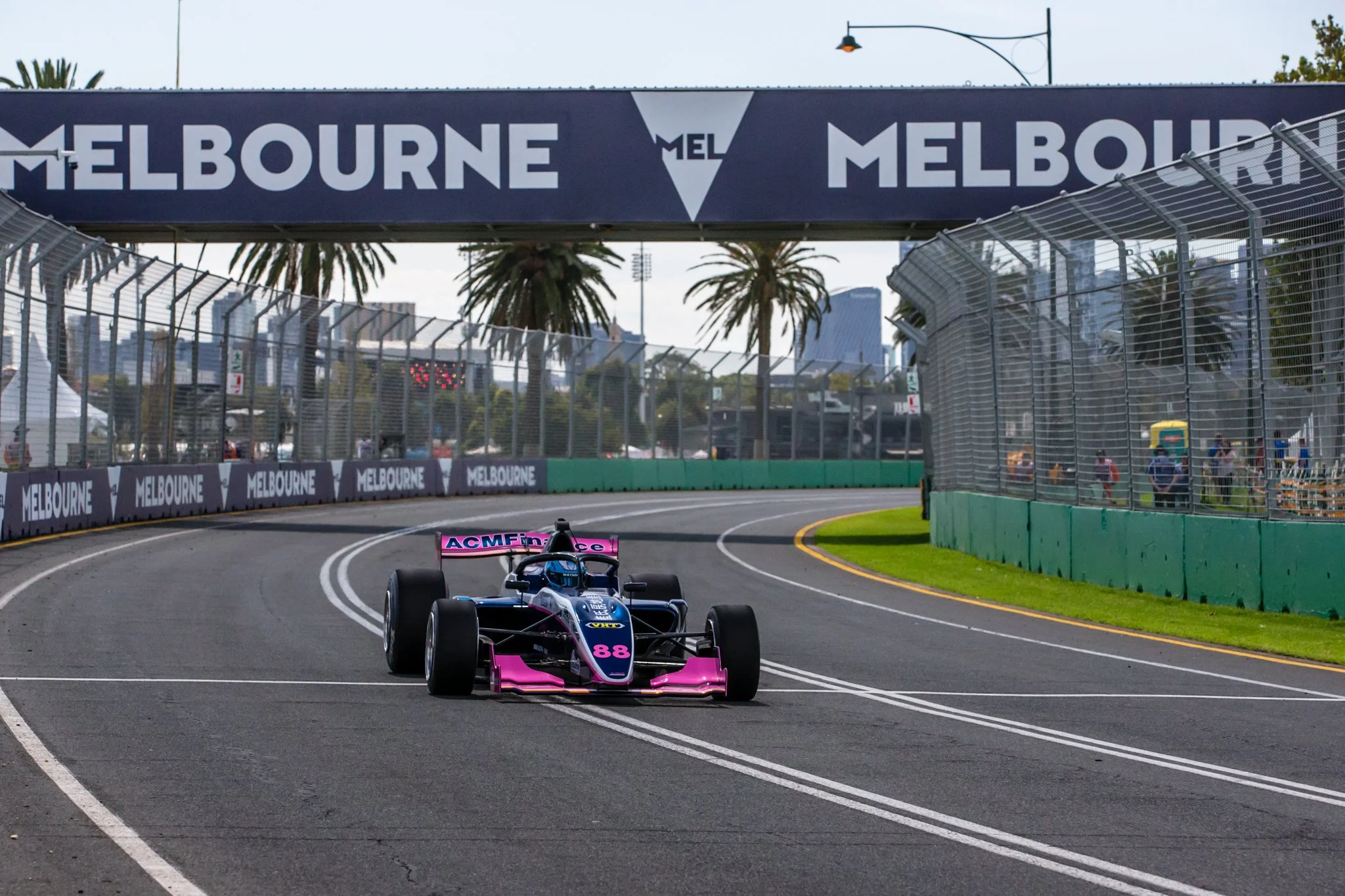 A Formula 1 race car with pink and black livery on a racing track, approaching a bridge with 'Melbourne' signage, and palm trees and city skyline in the background.