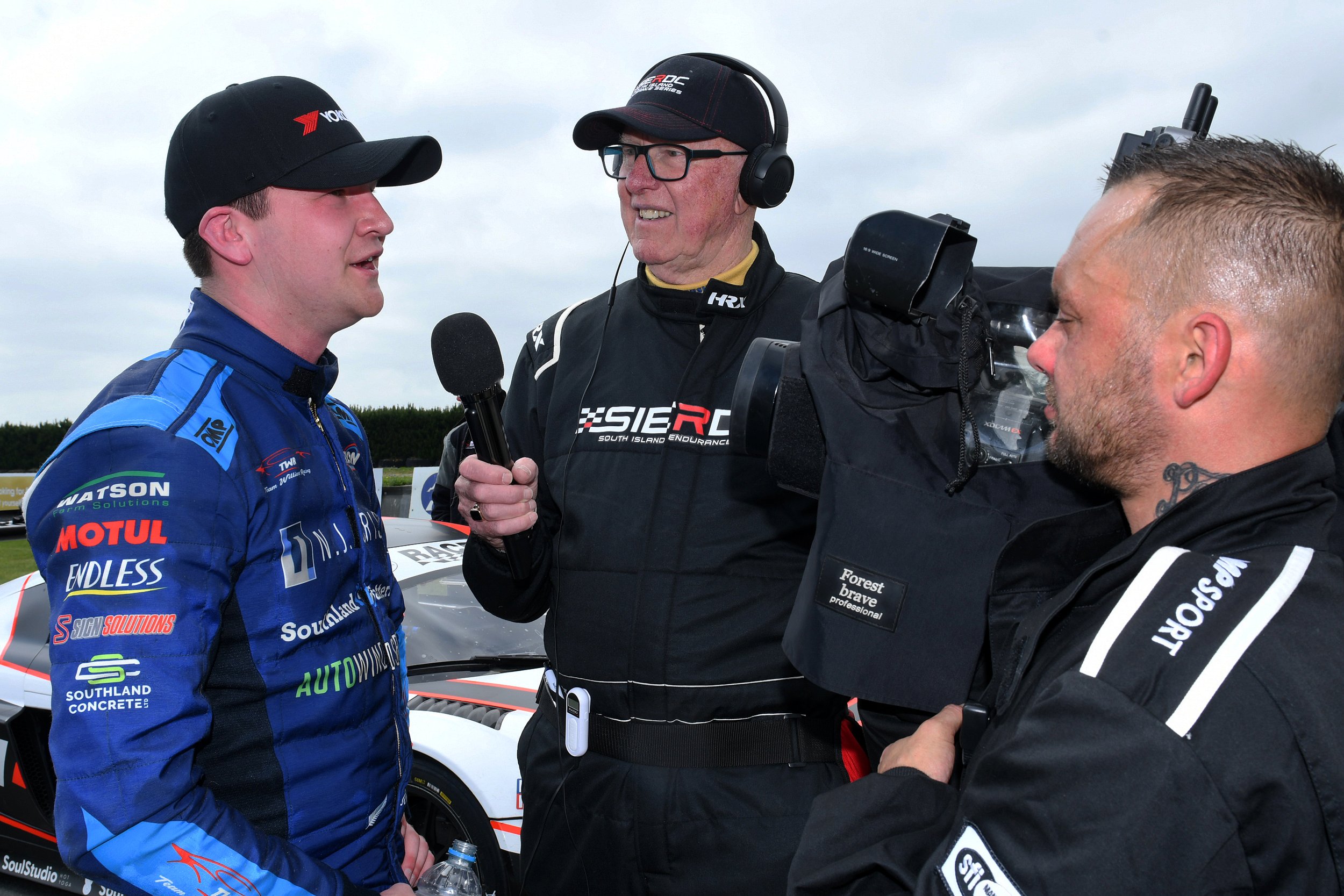A race car driver in a blue racing suit being interviewed by a man with glasses and a headset, while another crew member speaks. They are standing next to a racing car.