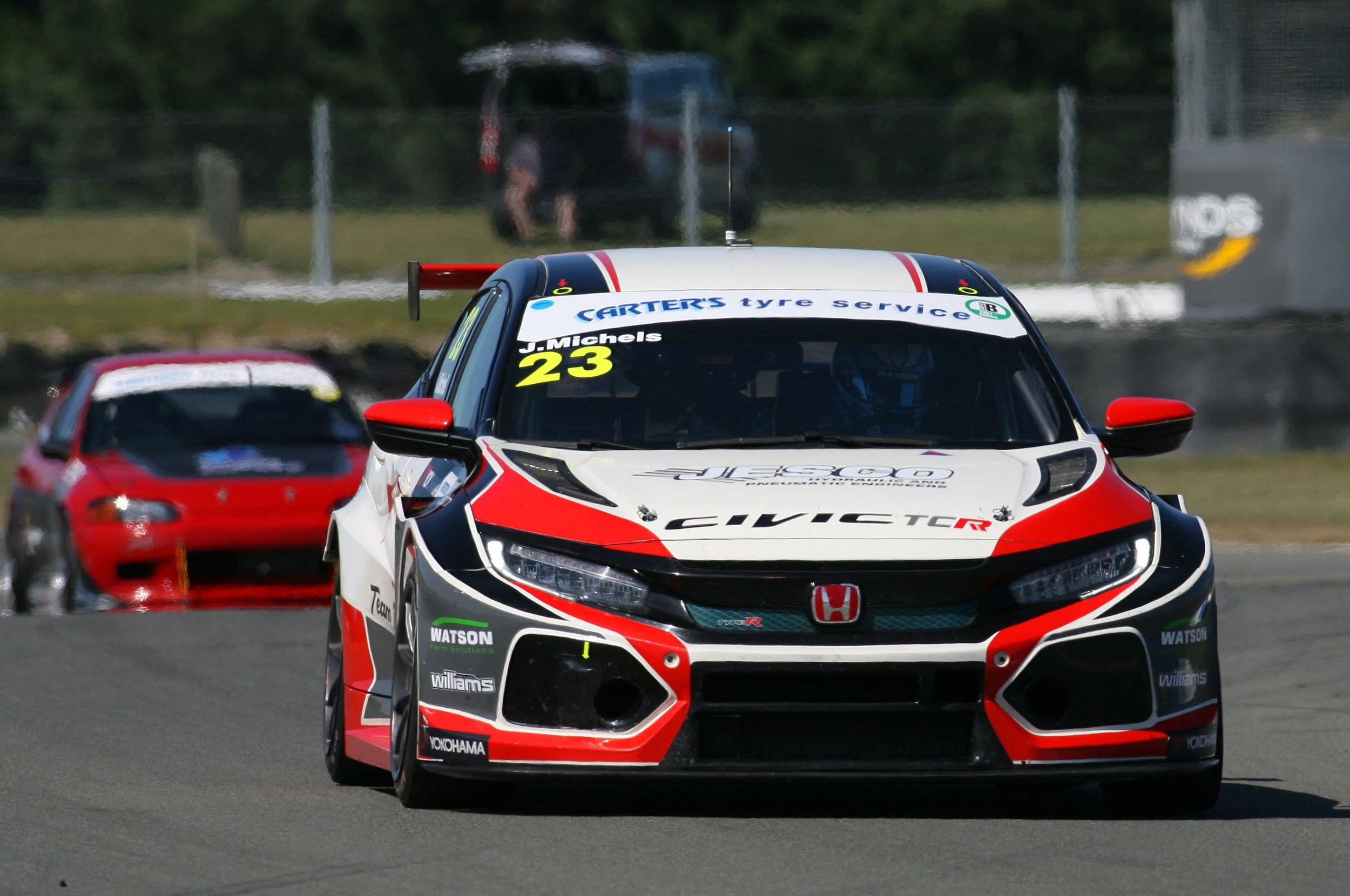 A white, red, and black Honda Civic Type R race car with the number 23 and driver J. Michels on the windshield, competing on a race track with a red car behind it and a blurred background of a person and a vehicle.